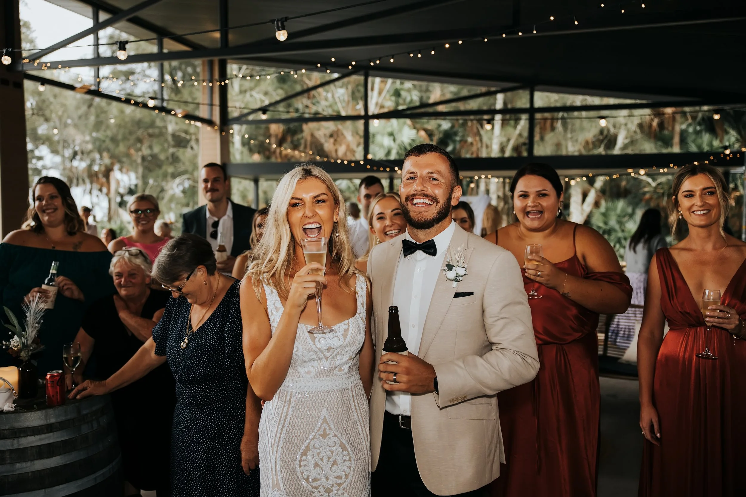 A wedding reception with guests, including a smiling bride and groom in formal attire, holding drinks, celebrating indoors decorated with string lights and greenery.