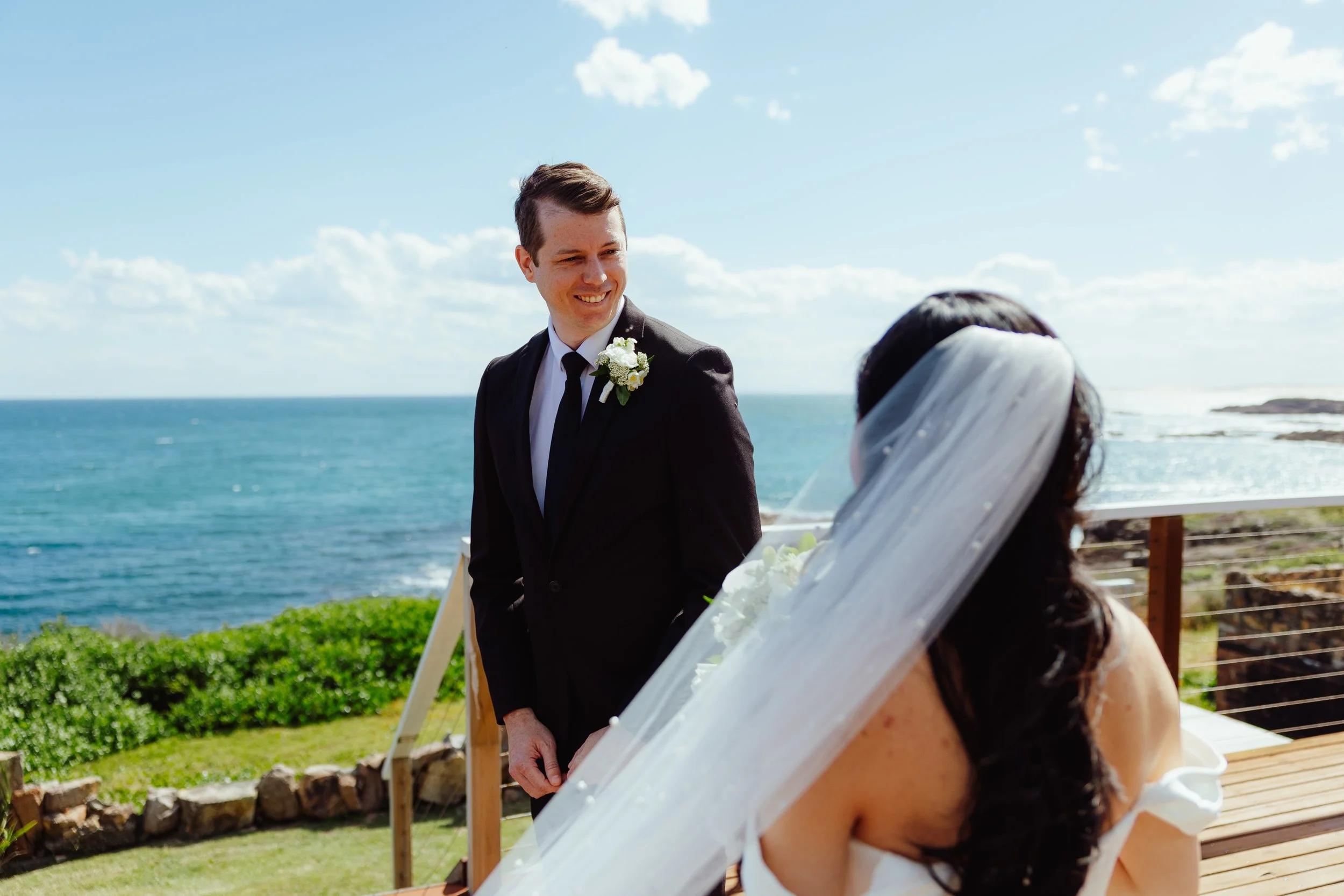 A wedding ceremony takes place outdoors by the ocean on a sunny day. The groom, in a black suit with a white shirt and black tie, is smiling at the bride. The bride, with long dark hair, is wearing a white dress and veil, and is seated on a bench.