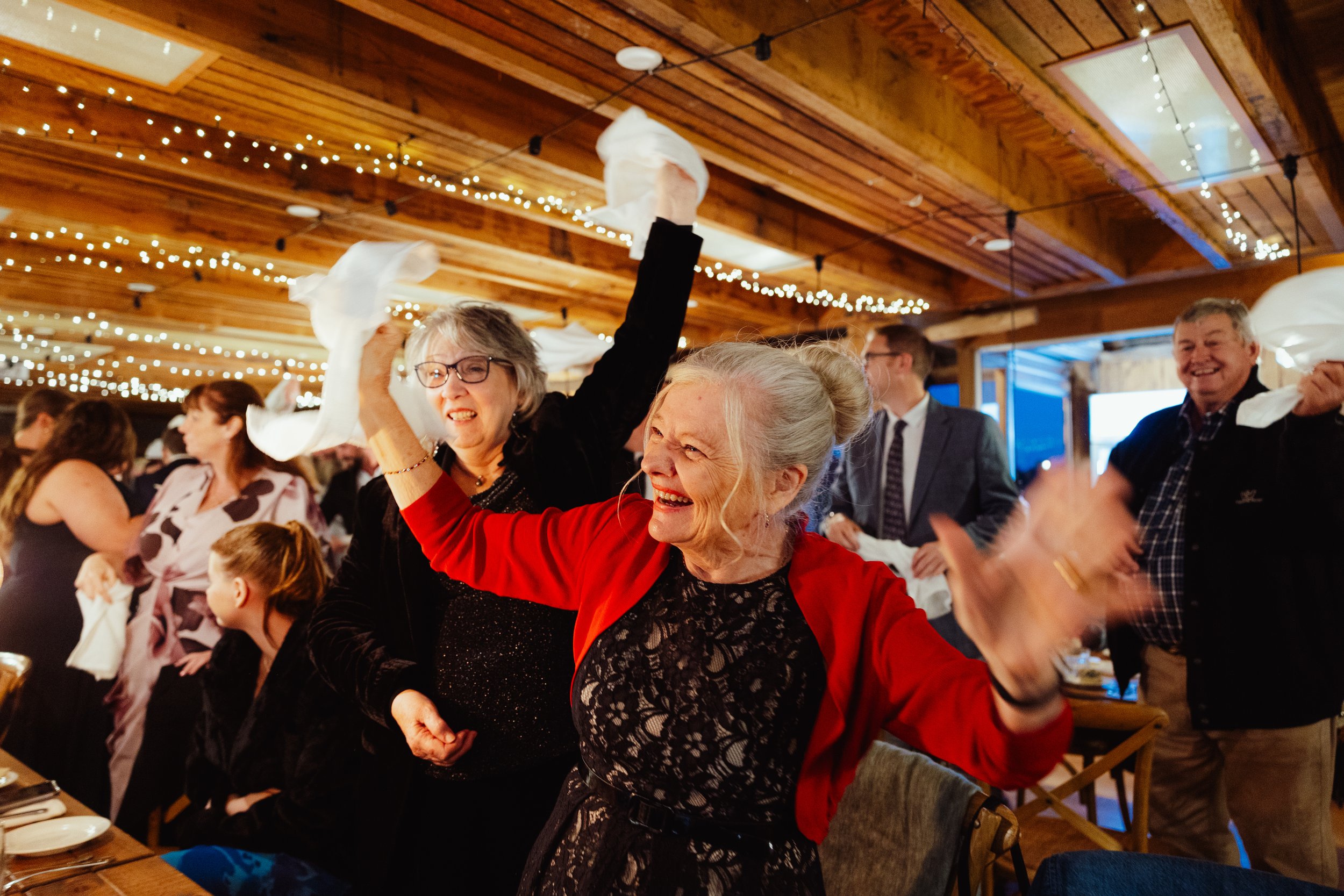 People celebrating at a festive gathering with string lights on the wooden ceiling, some holding napkins in the air, smiling and having fun.