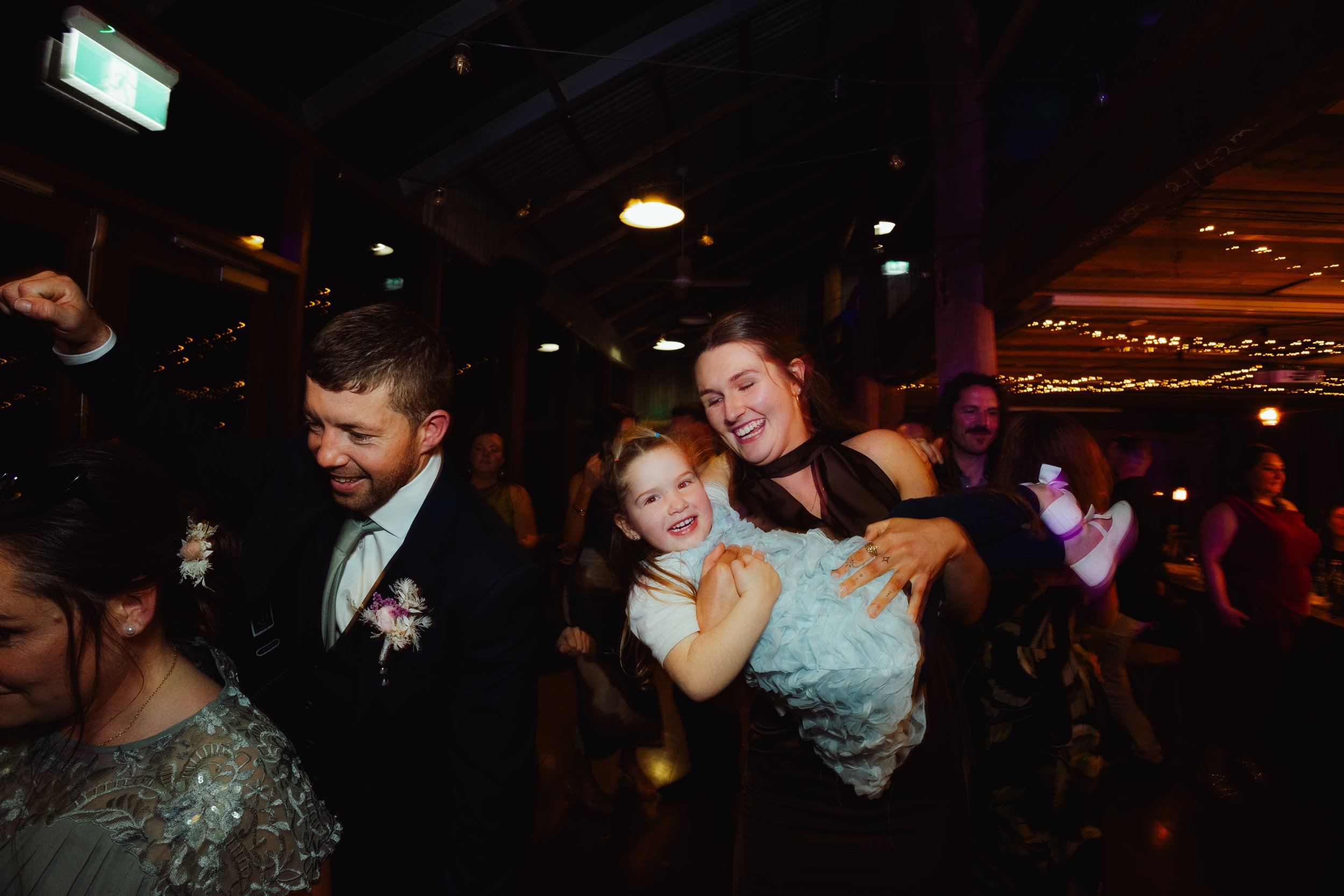People celebrating at a party or wedding reception, dancing and smiling, with string lights and wooden ceiling visible.