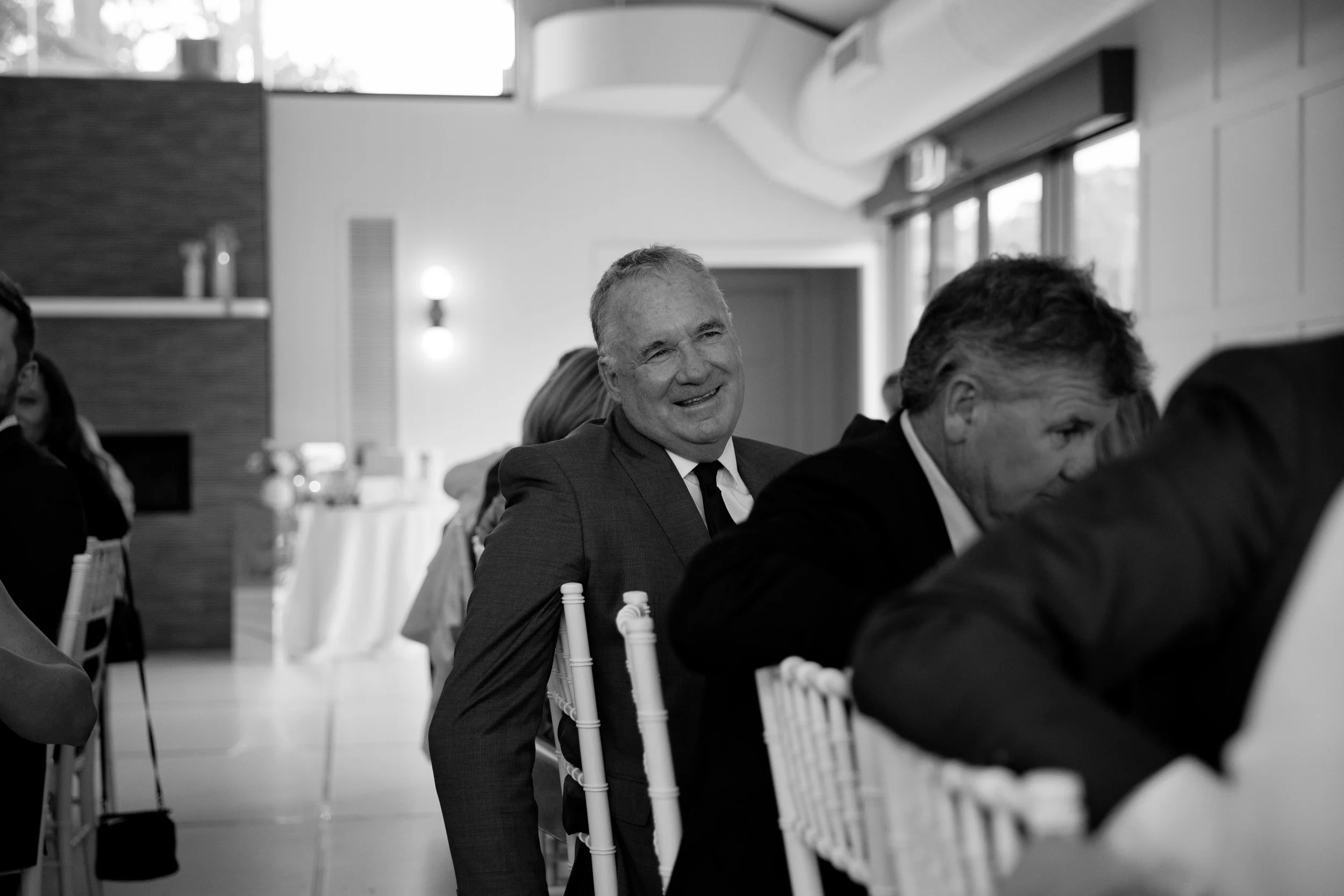 A black and white photo of a group of people seated at a round table during a formal event in a bright room with high ceilings. The man in the center, smiling and wearing a suit and tie, is facing the camera. Other people are partially visible around
