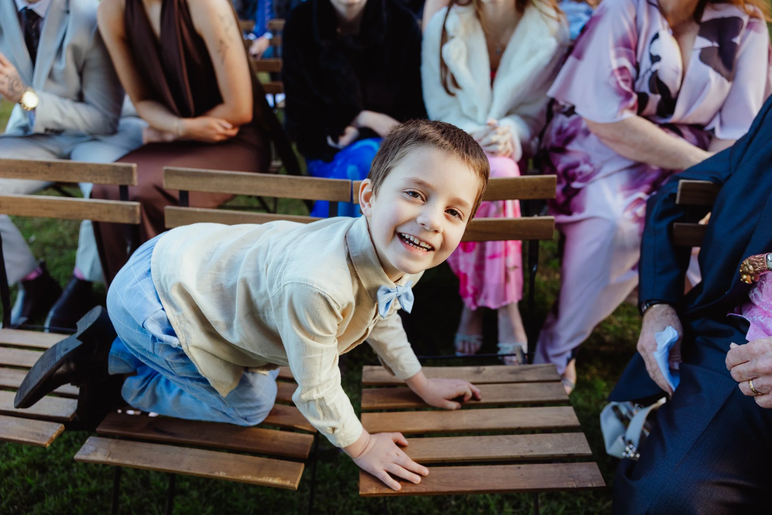 A young boy smiling and leaning on a wooden bench at an outdoor event, with several adults sitting behind him.