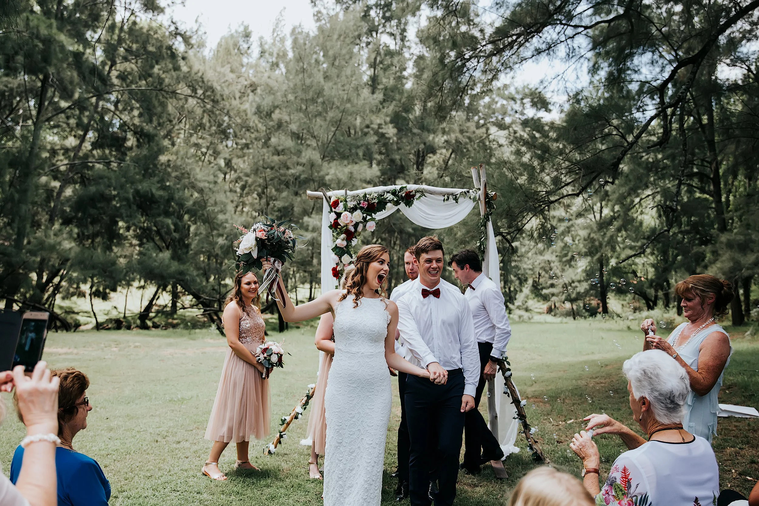 A wedding ceremony taking place outdoors in a park with guests sitting on the grass, a bride and groom holding hands under a floral arch, surrounded by family and friends with trees in the background.