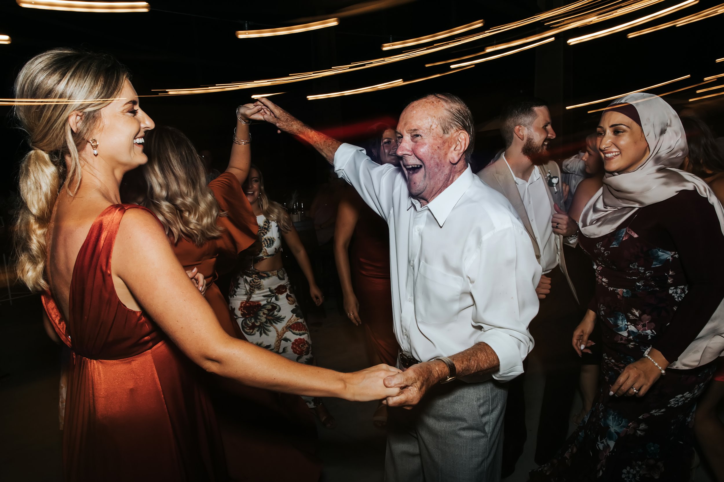 An elderly man and a woman are dancing together, holding hands and smiling at each other in a lively party with other people dancing in the background.