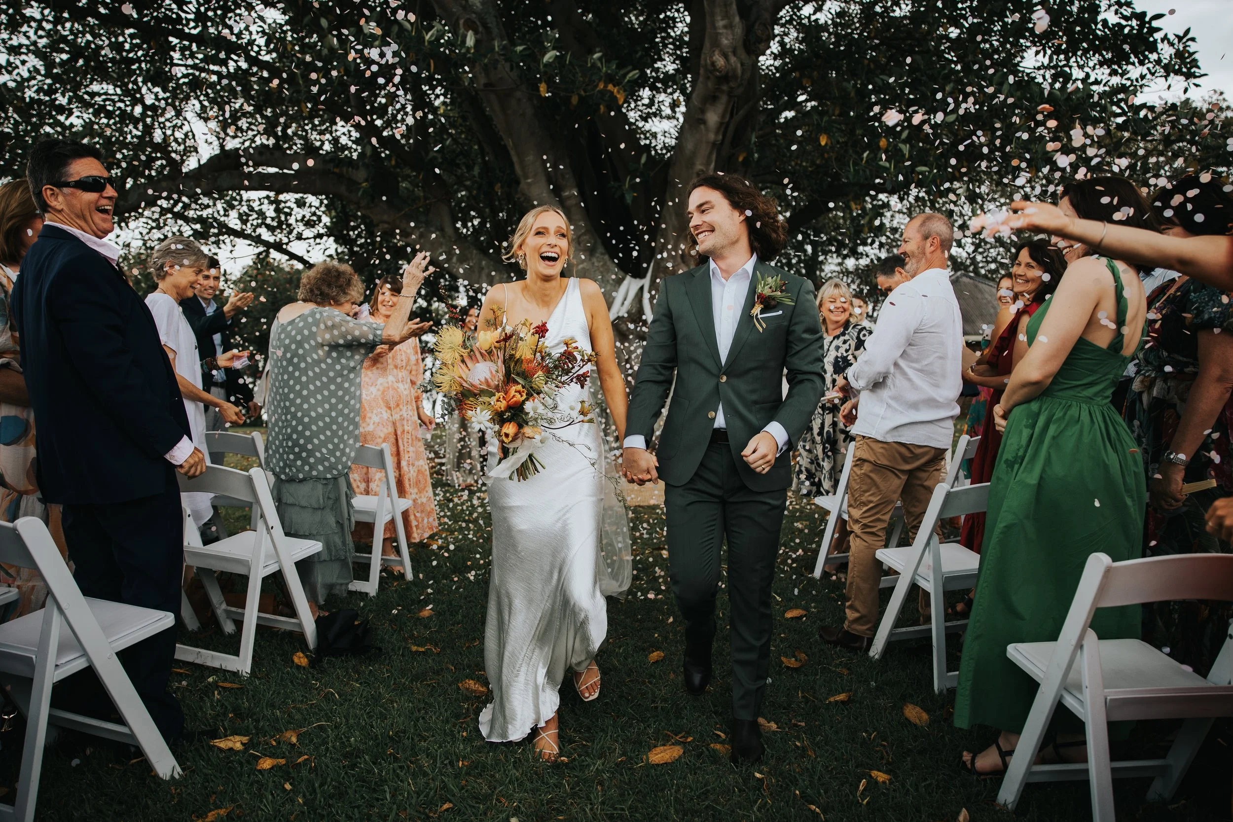 A bride and groom walk down the aisle at an outdoor wedding ceremony, surrounded by friends and family, with confetti falling from above, beneath a large tree.