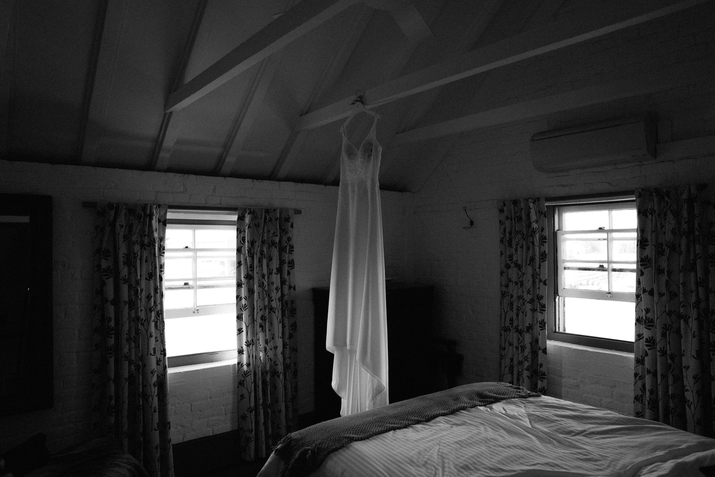 A bedroom with two windows with patterned curtains, a bed with a white comforter and dark blanket, and a dress hanging from the ceiling fan.