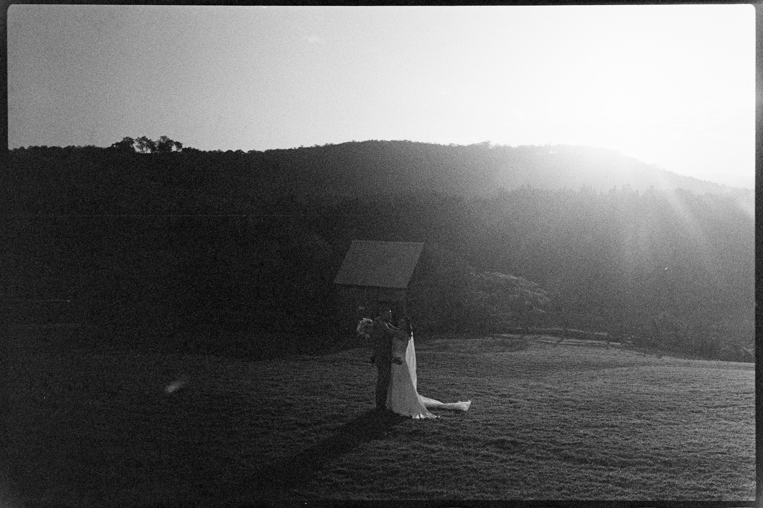 A black and white photo of a person in a swimsuit with long hair, bending over in a grassy open area near trees and hills, with sunlight illuminating the scene from the background.