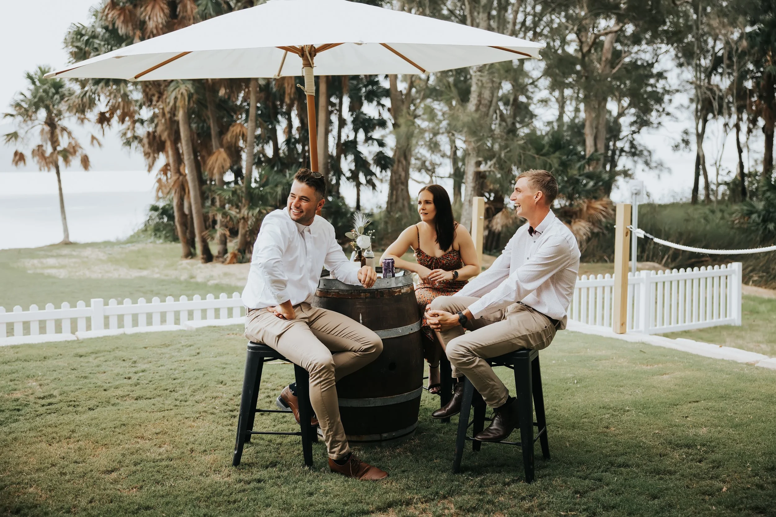 Three people sitting around a barrel table outdoors under a large white umbrella, with trees and a grassy area in the background, smiling and talking.