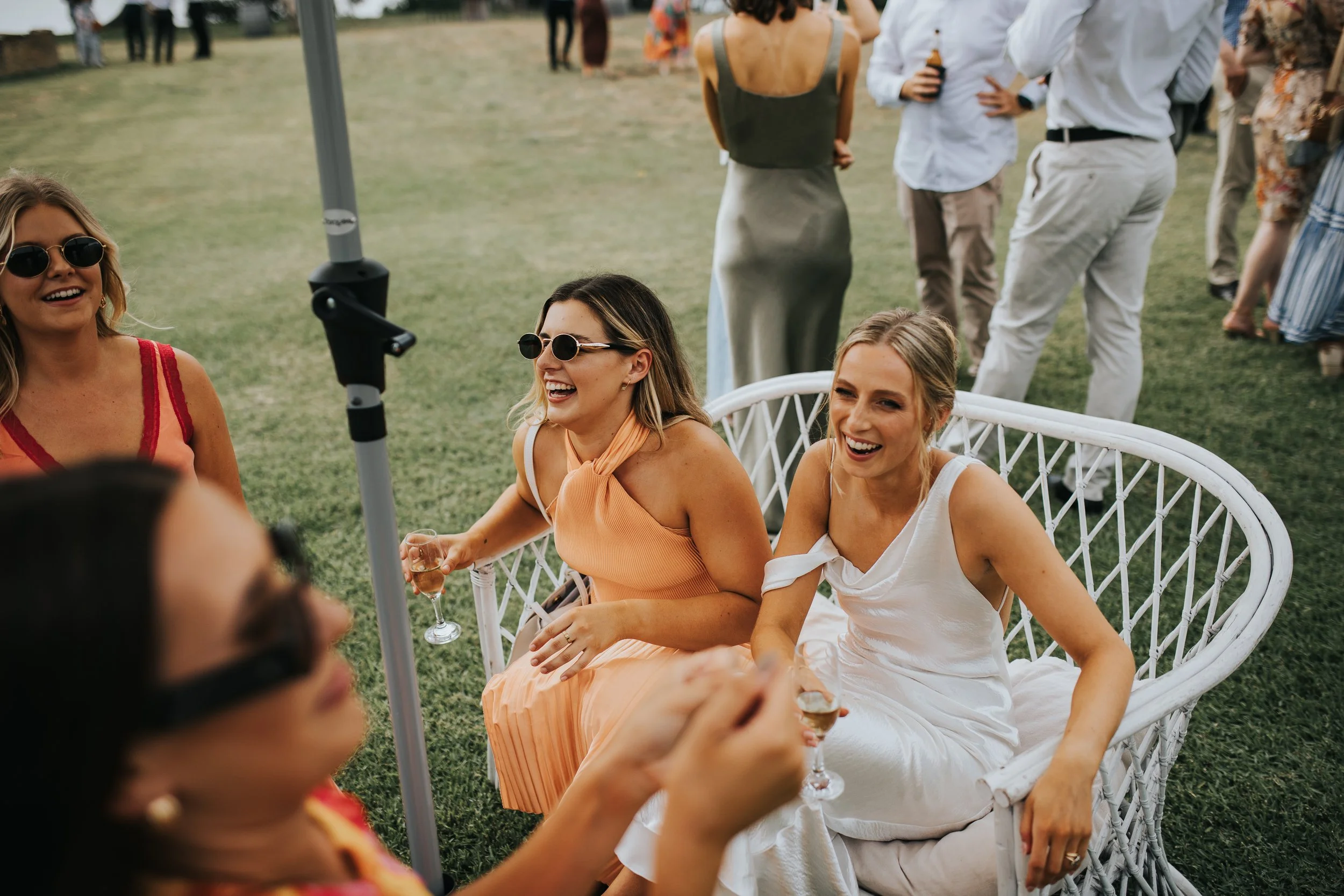 A group of women sitting on a white wicker chair outdoors, laughing and holding glasses of champagne, with more people standing and chatting in the background at a sunny outdoor gathering.