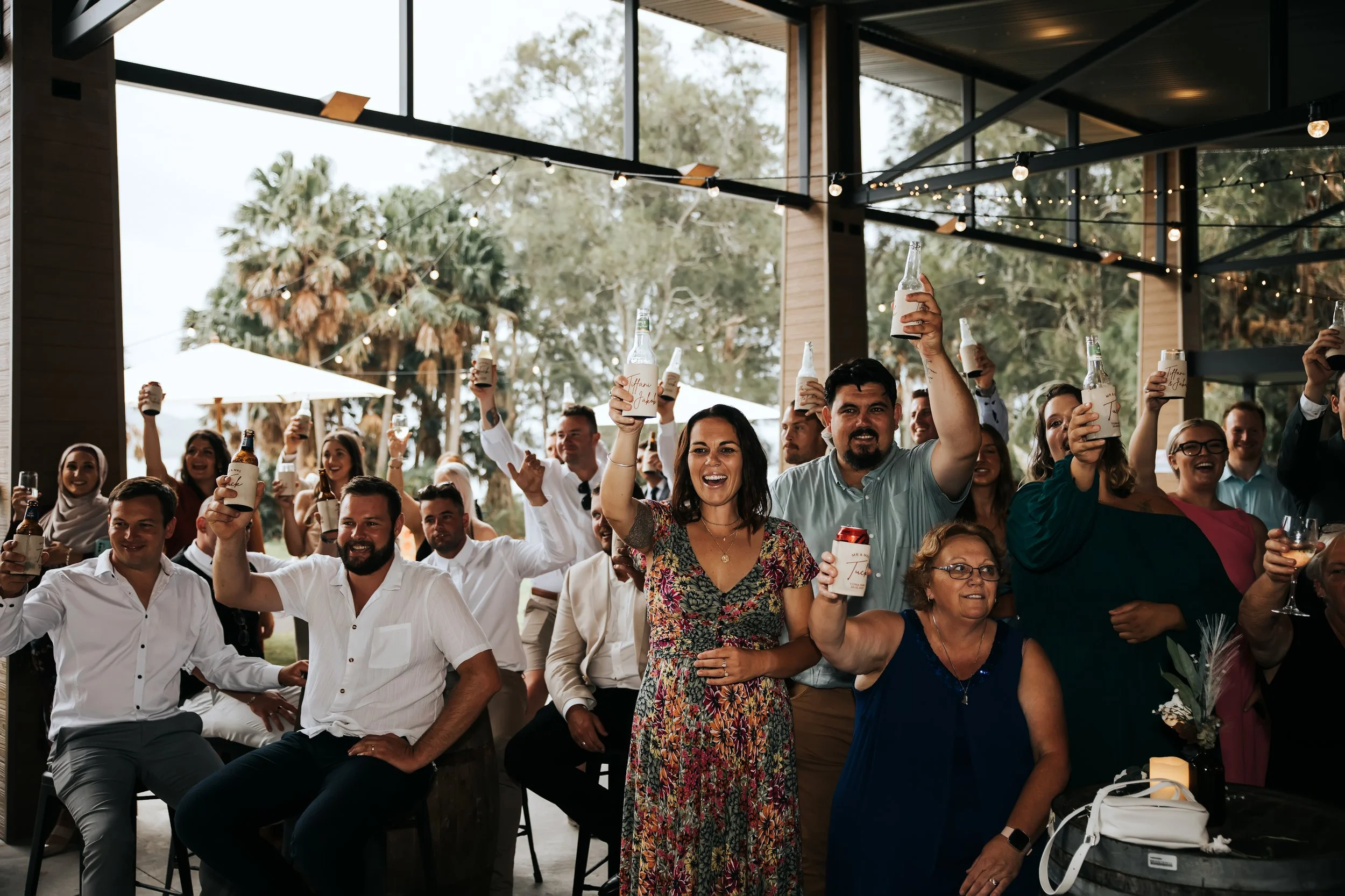People gathered indoors at a celebration, raising bottles and glasses, smiling and enjoying the event.