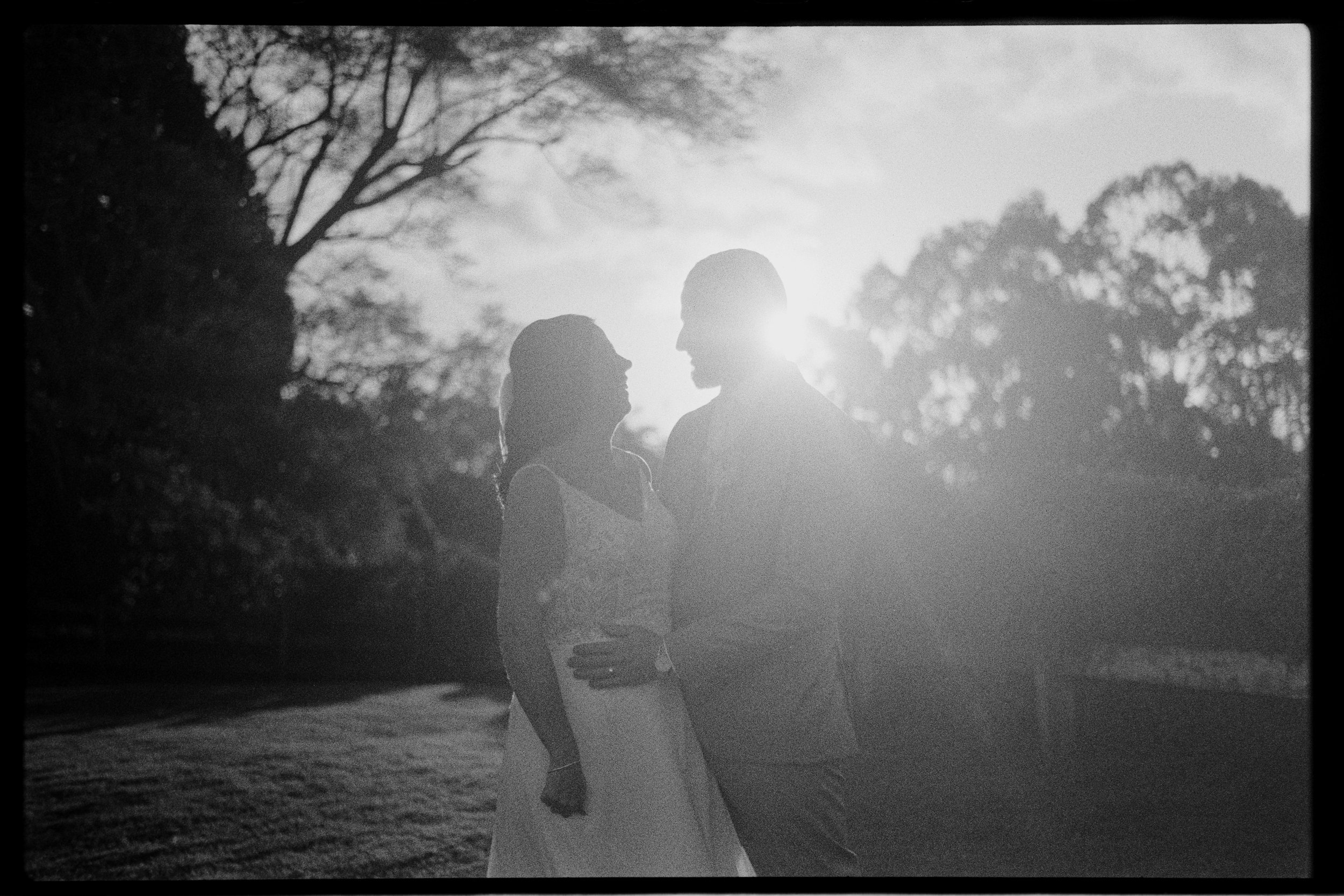 A black and white photo of a couple standing close together outdoors, facing each other, with the sun shining behind them. The scene includes trees and a natural setting.