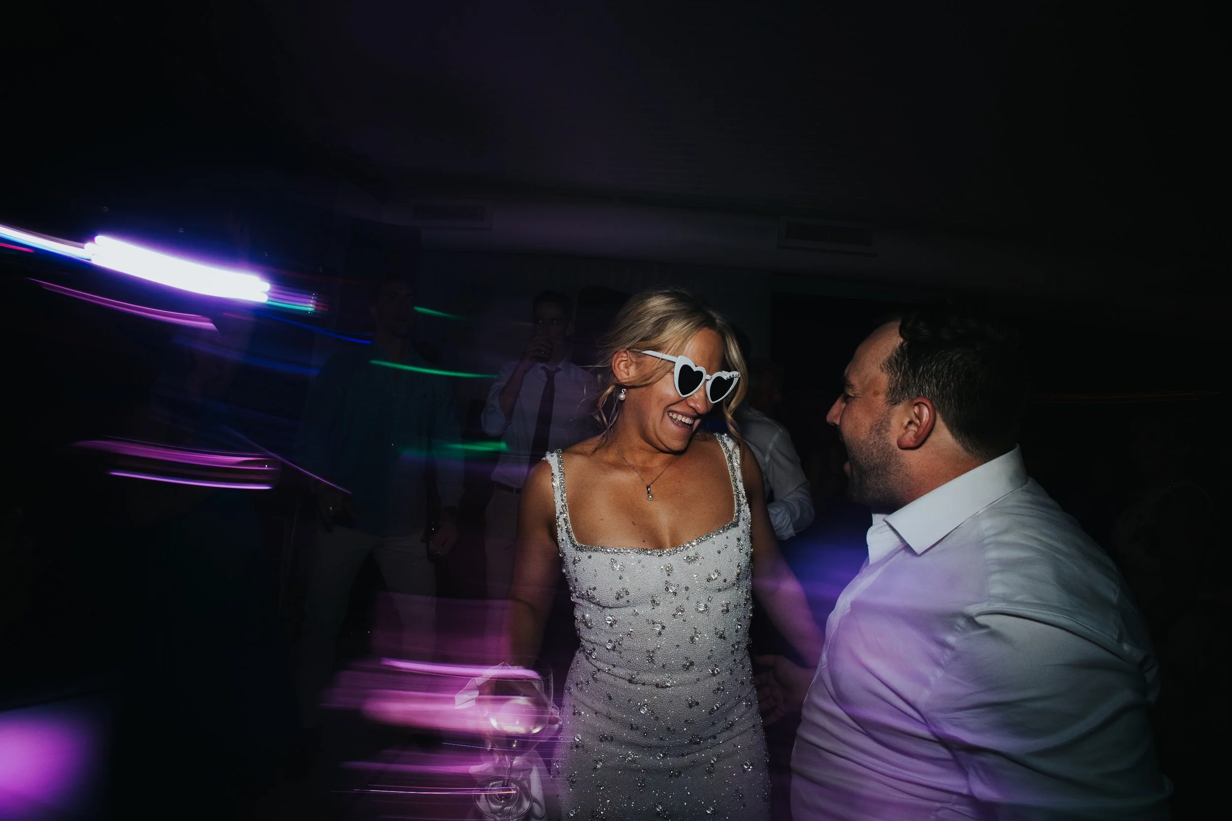 A woman with blonde hair wearing heart-shaped sunglasses and a dress with sequins, smiling and dancing with a man in a white shirt at a party with colorful lights.
