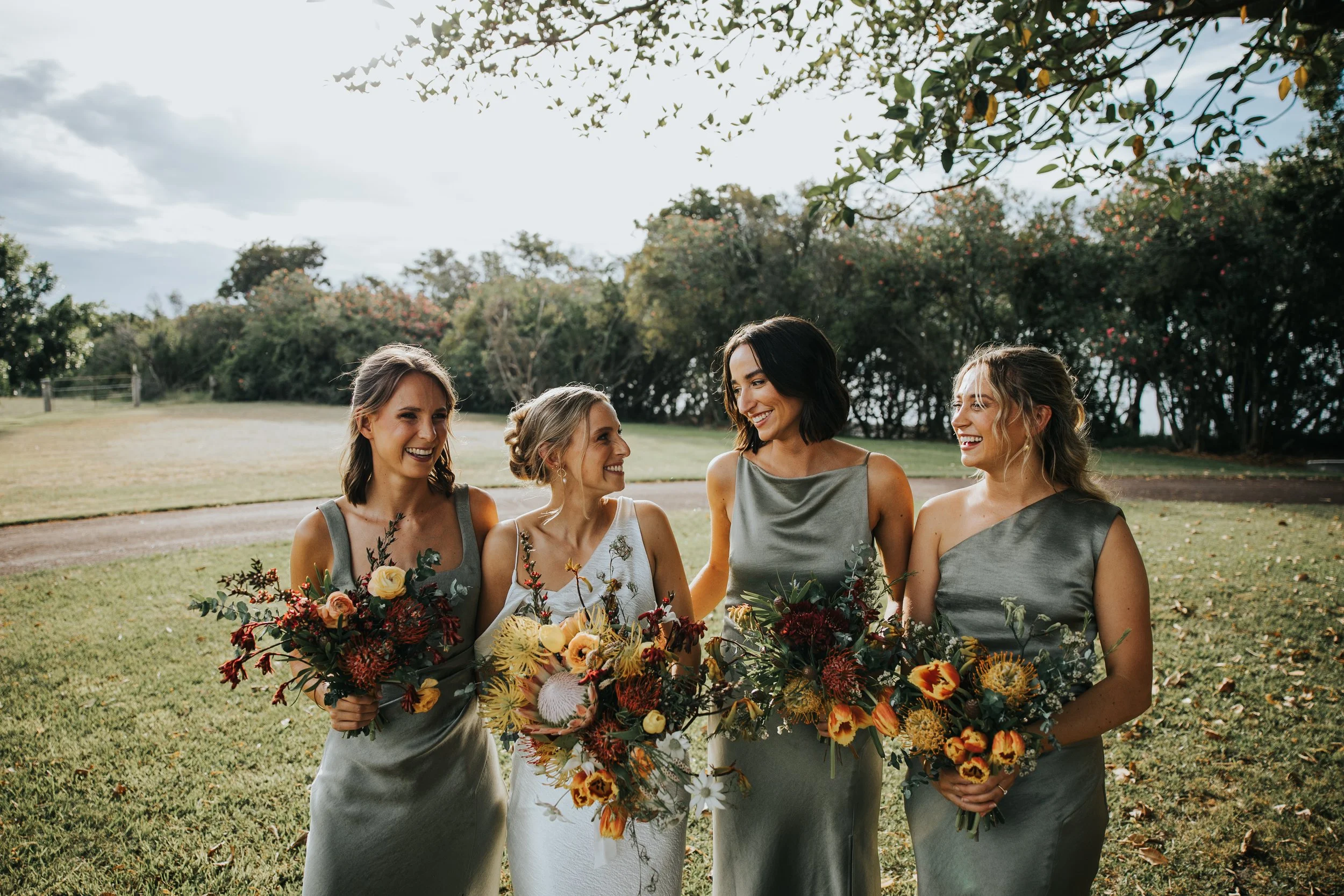 A group of four women in gray dresses, standing outdoors on a grassy field, smiling and holding large bouquets of colorful flowers, with trees and cloudy sky in the background.