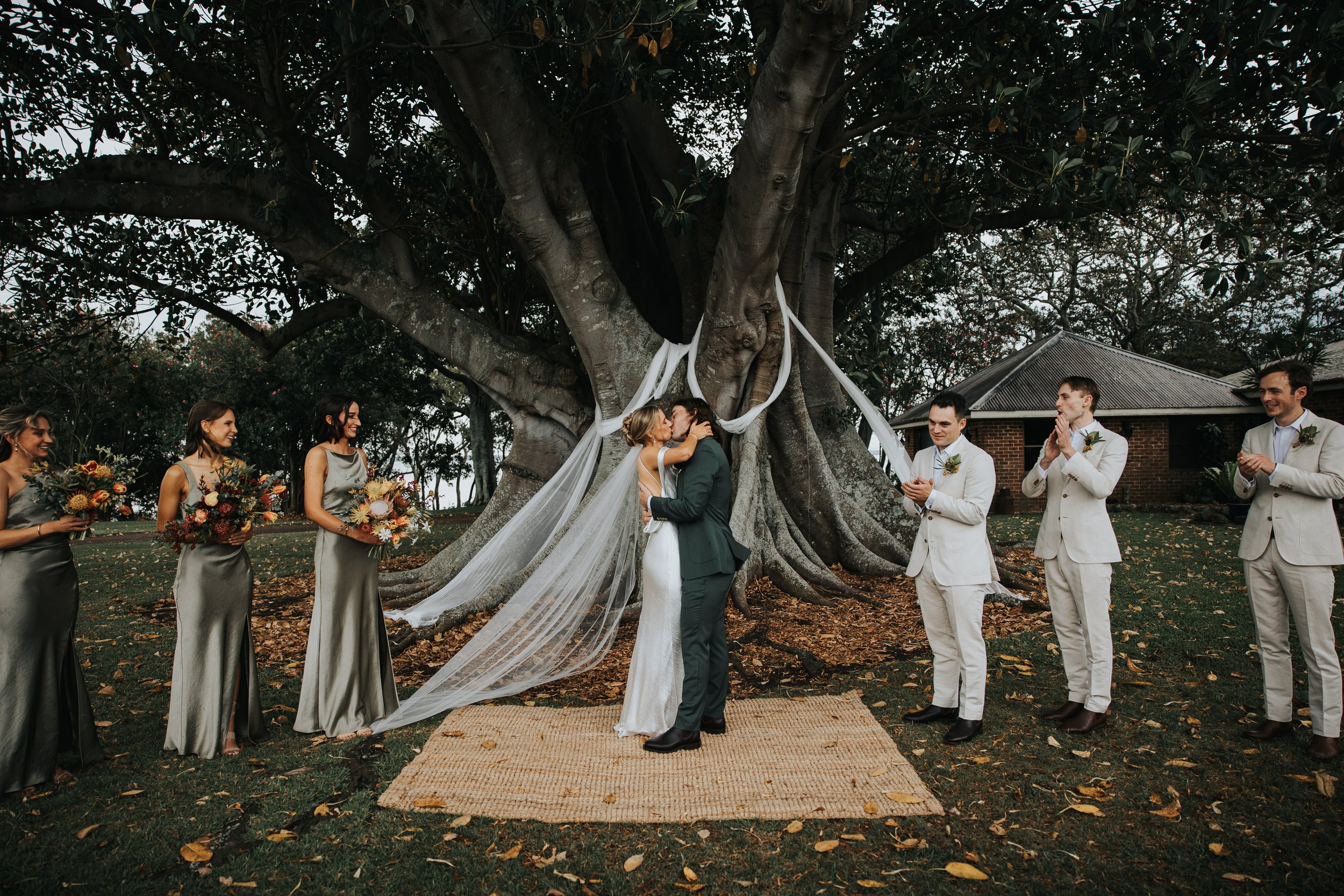 A wedding ceremony outdoors under a large tree, with a couple kissing in front of friends and family. The bride is in a white gown with a long veil, and the groom is in a dark suit. Bridesmaids wear matching gray dresses holding bouquets, and groomsm