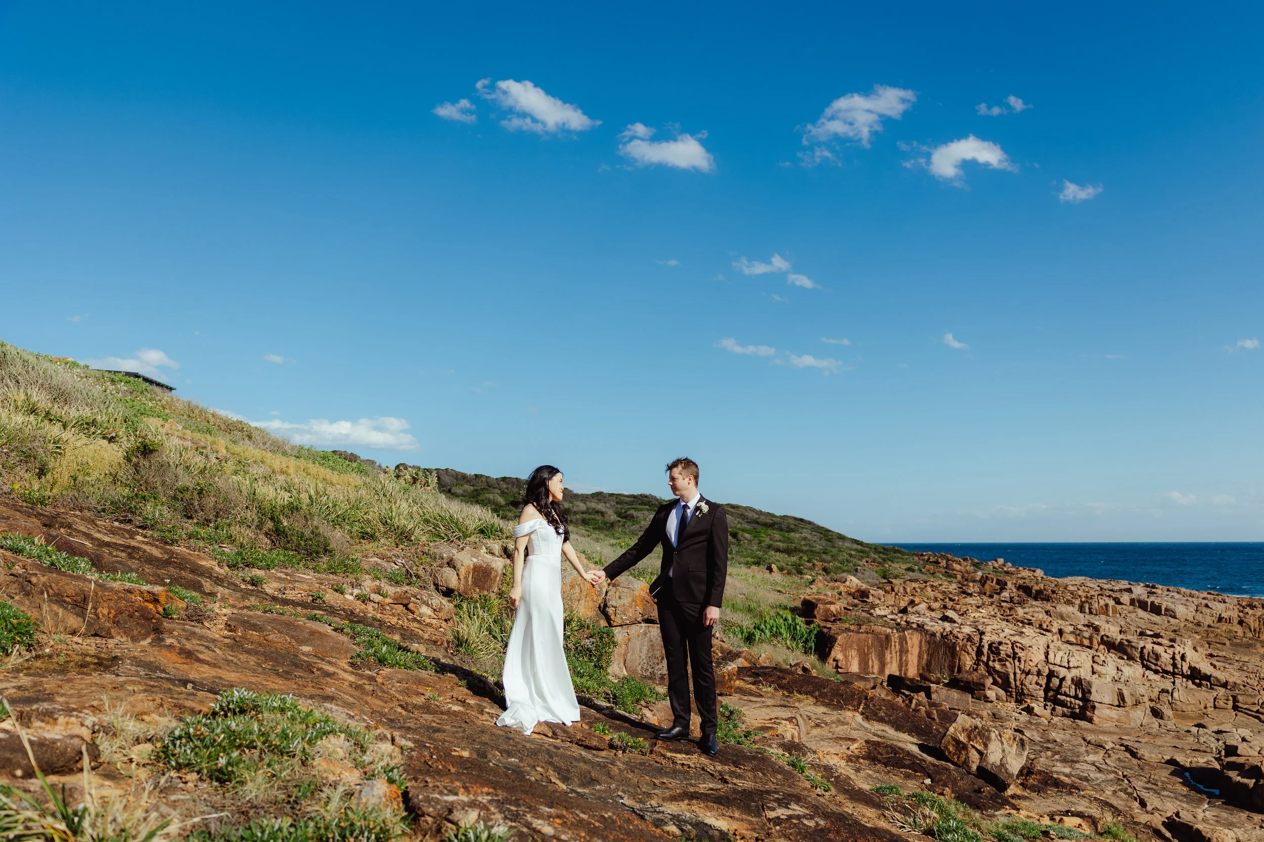 A couple dressed in wedding attire holding hands on a rocky hillside near the ocean under a blue sky with some clouds.