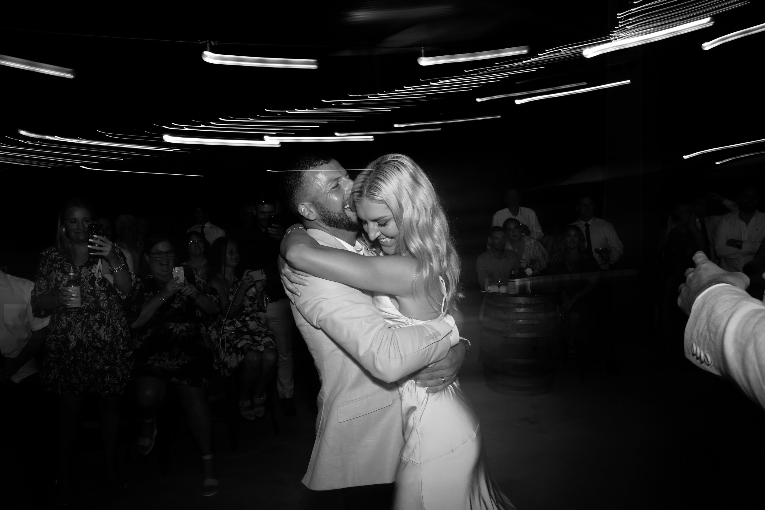 A joyful couple dances closely together at a party or wedding reception, surrounded by guests watching and taking photos, with ceiling lights creating motion blur in the background.
