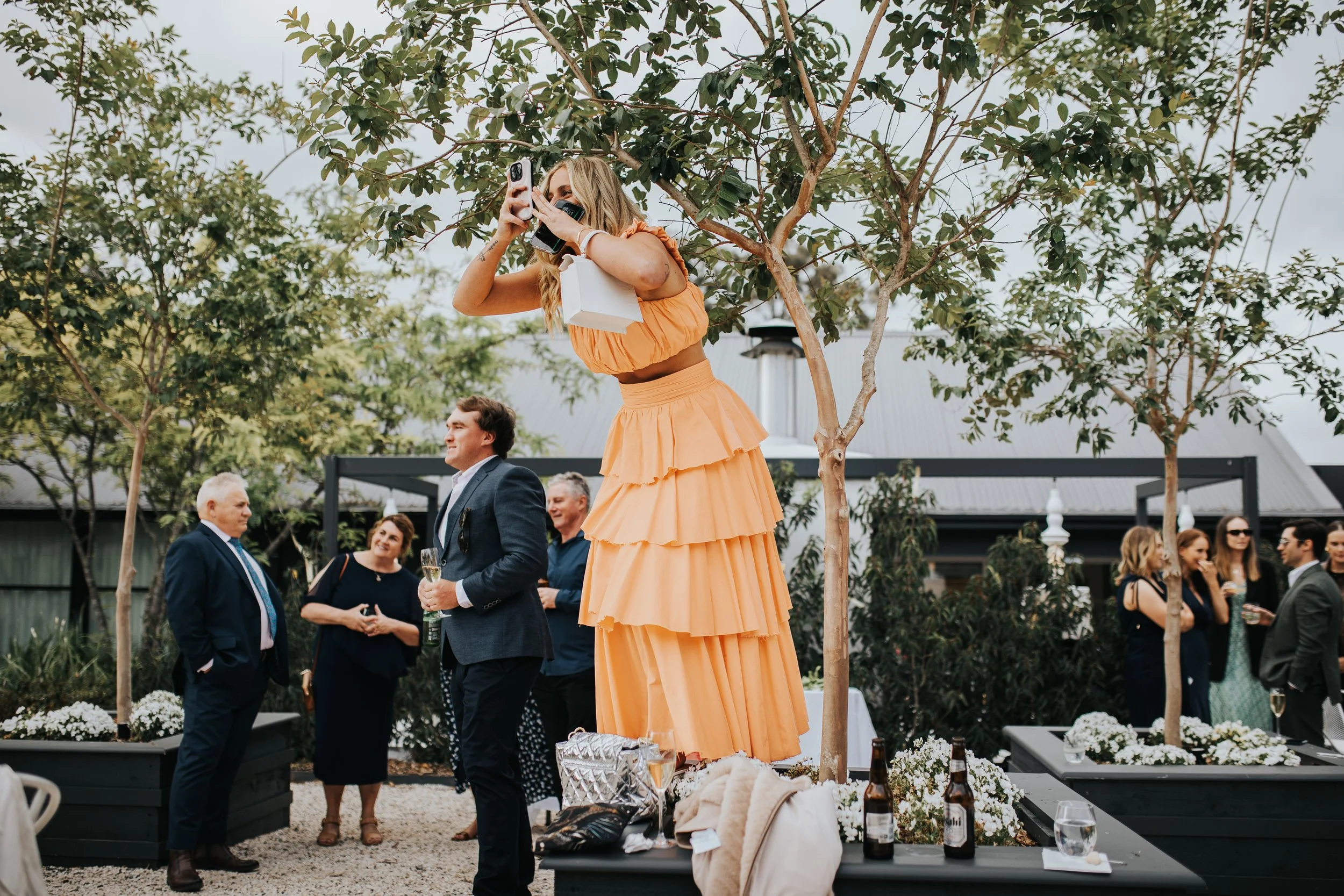 A woman in an orange dress speaking into a microphone at an outdoor event, surrounded by people socializing and holding drinks, with trees and a building in the background.