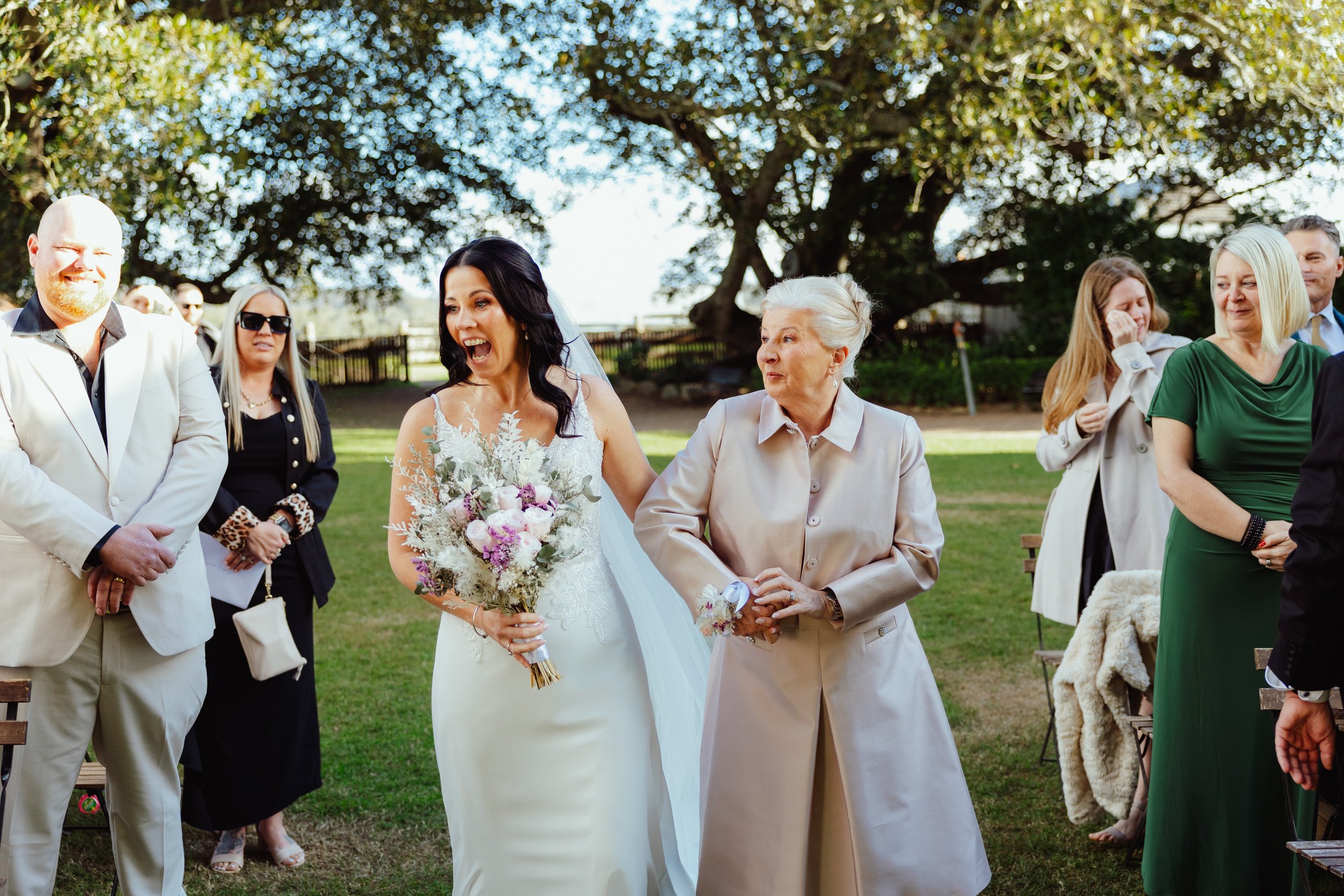 A bride holding a bouquet, smiling, standing next to an older woman, with people gathered outdoors under a large tree during a wedding ceremony.