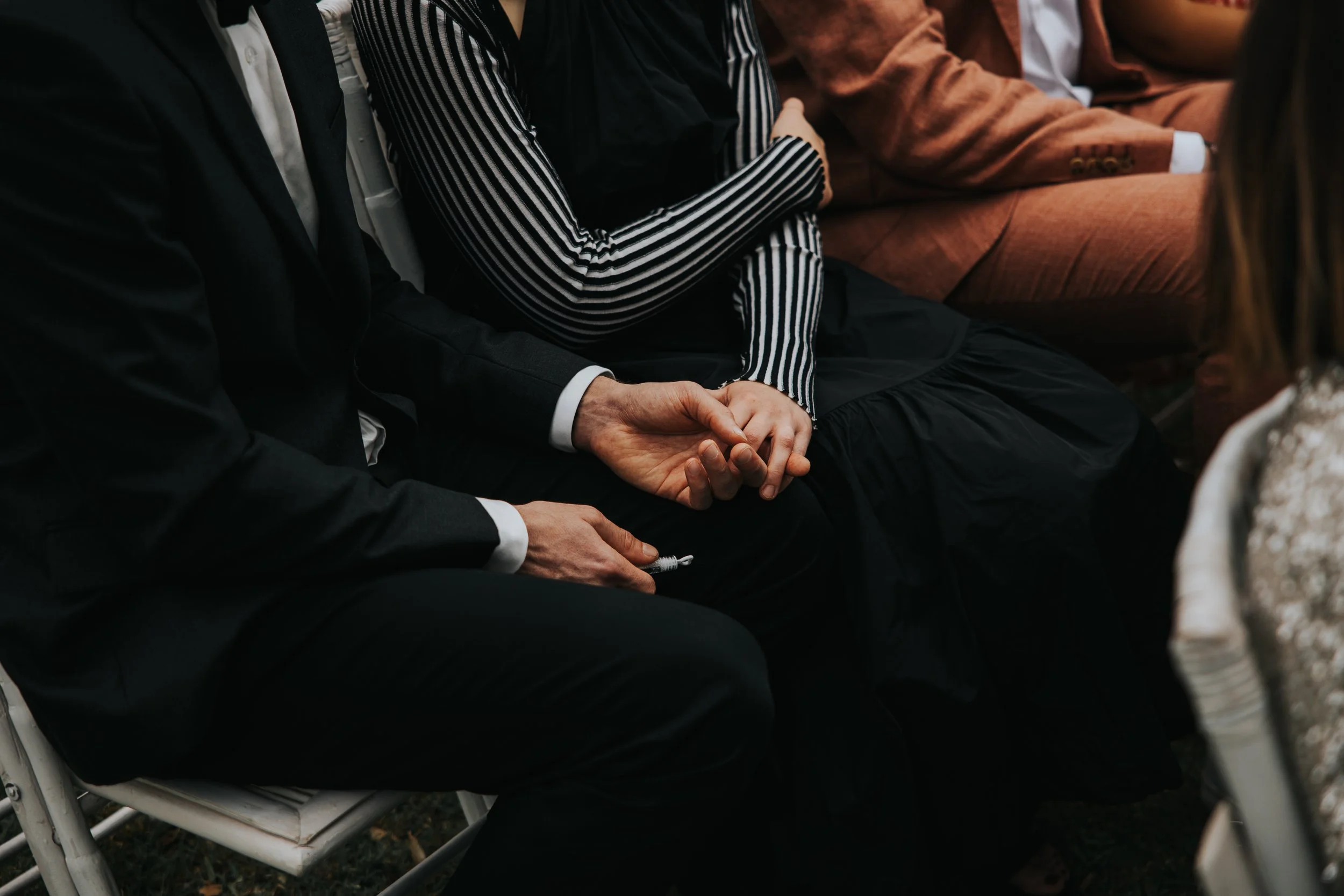 Close-up of people seated in a row, holding hands, dressed in formal and semi-formal attire, with some wearing suits and one person in a striped shirt, sitting on white chairs.