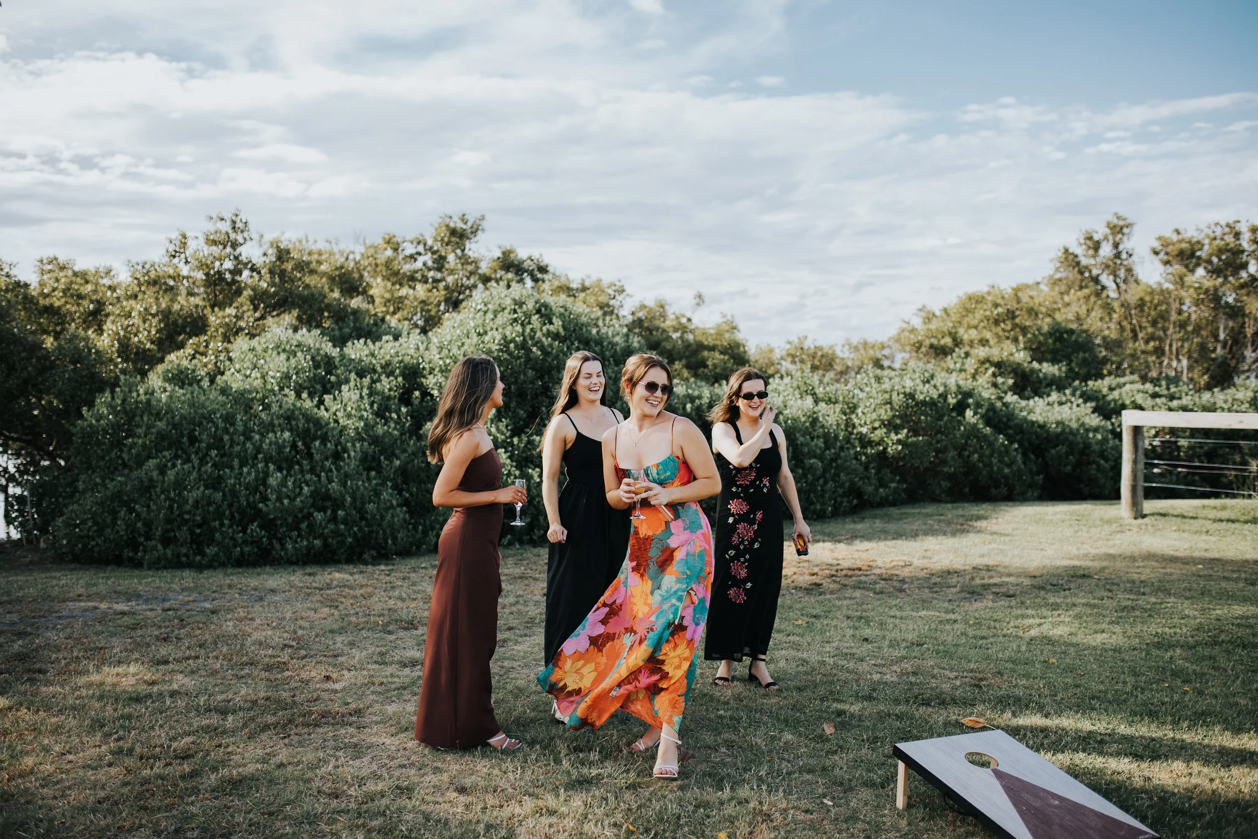 Four women in summer dresses and sunglasses walking and talking outdoors on a sunny day, with trees and a partly cloudy sky in the background.