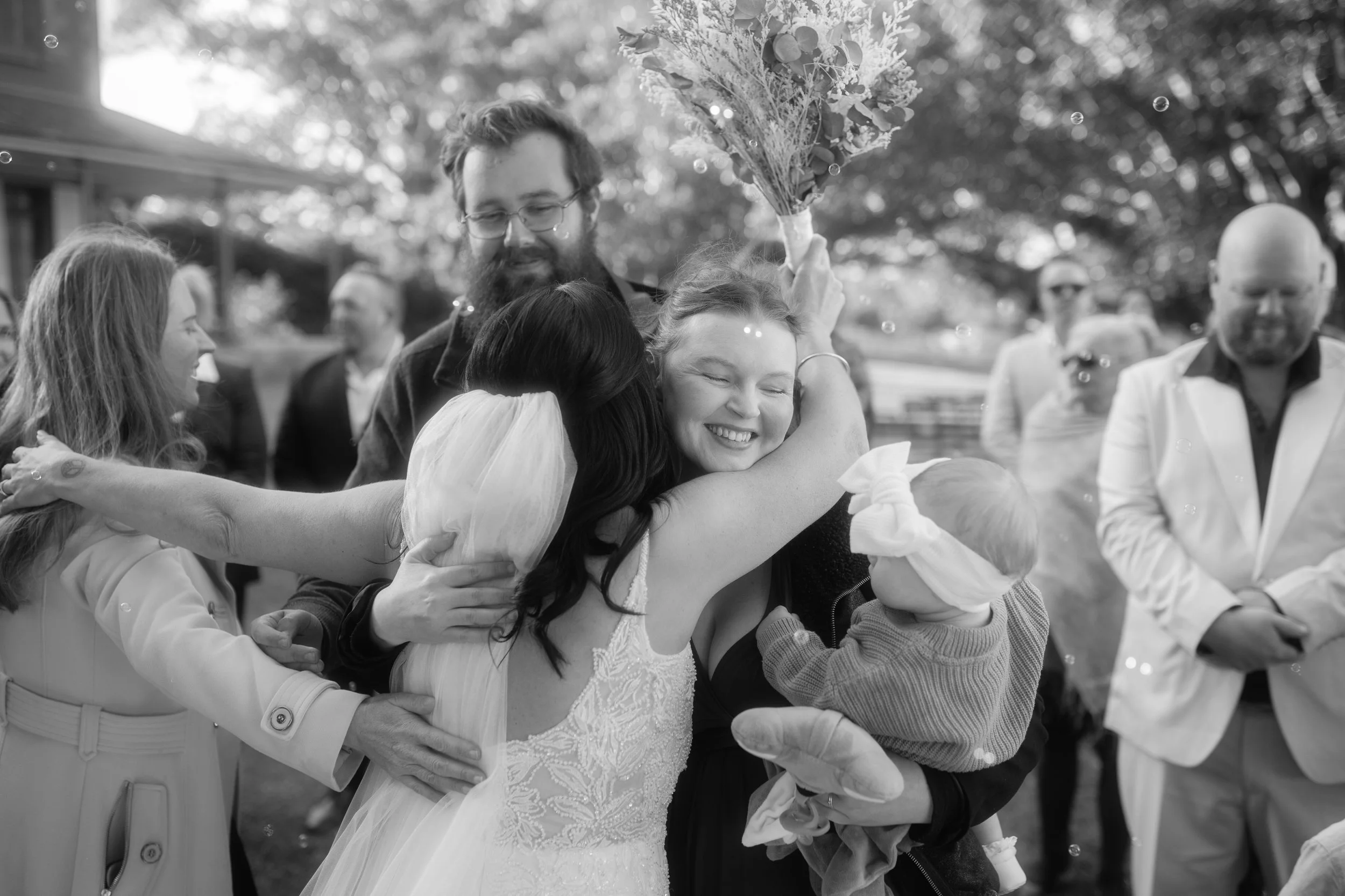A group of people are hugging each other in an outdoor setting, with a woman smiling while holding a bouquet of flowers.