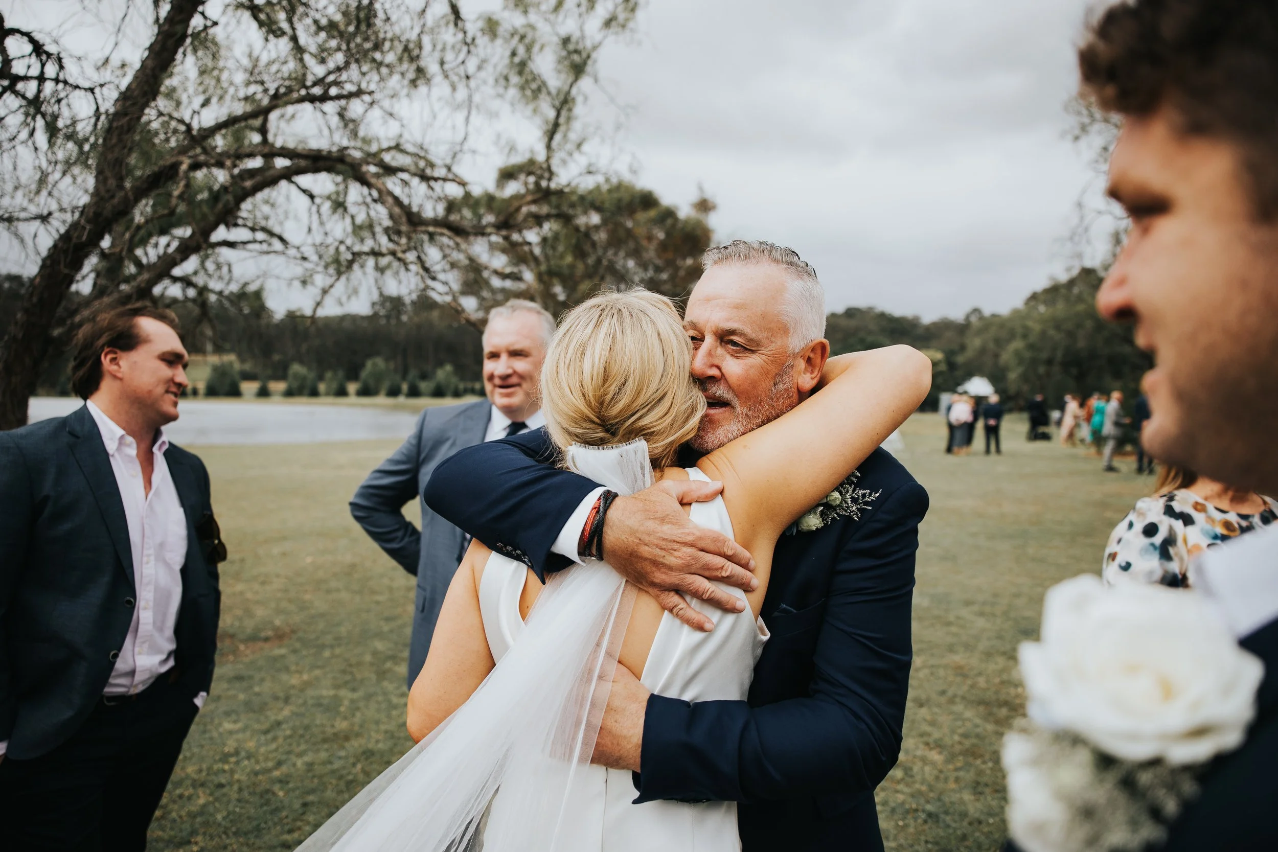 A group of people outdoors, with one couple hugging and smiling, dressed in formal attire, in a scenic park with trees and a grassy area.