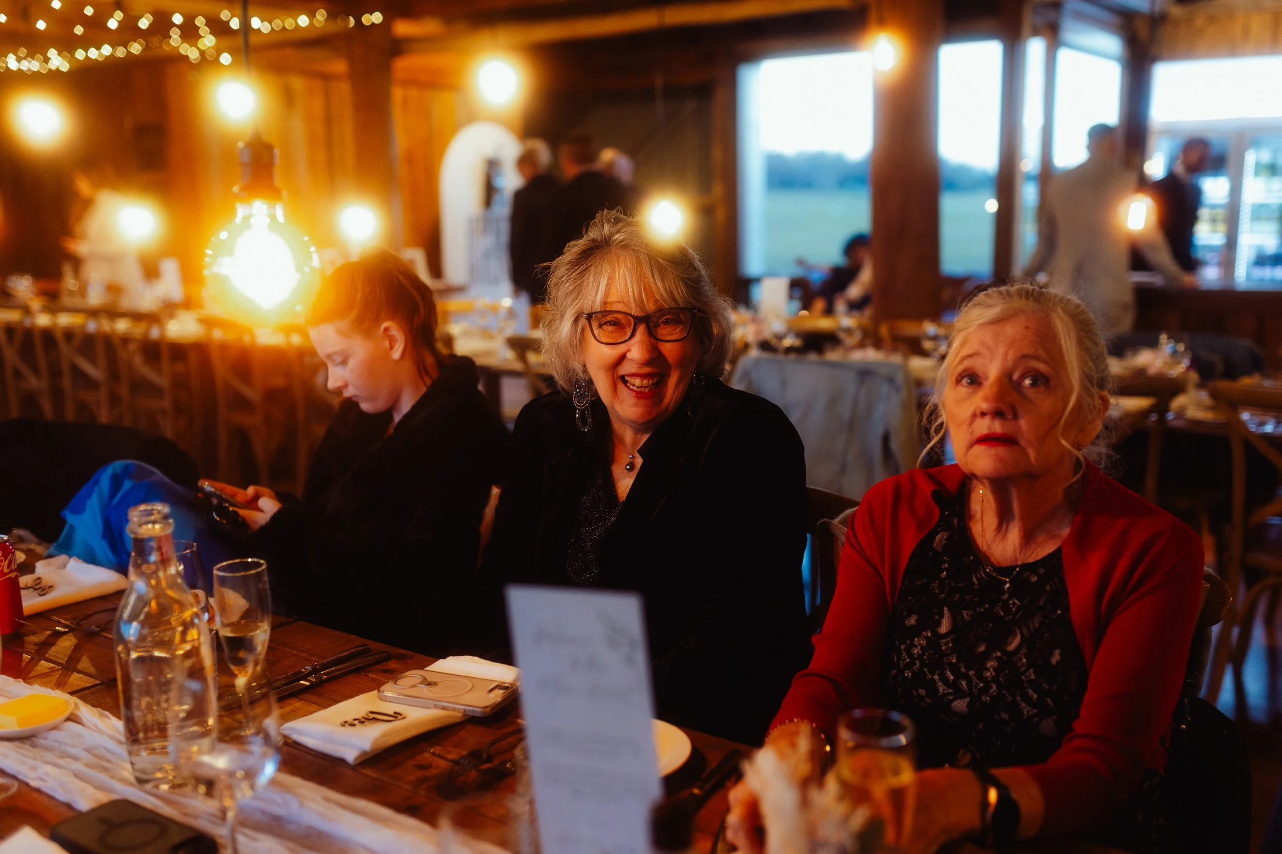 People gathered around a table at a warm, rustic wooden restaurant with string lights on the ceiling. There are three women in the foreground, two older women smiling at the camera and a younger woman looking down at her phone. The table has plates, 