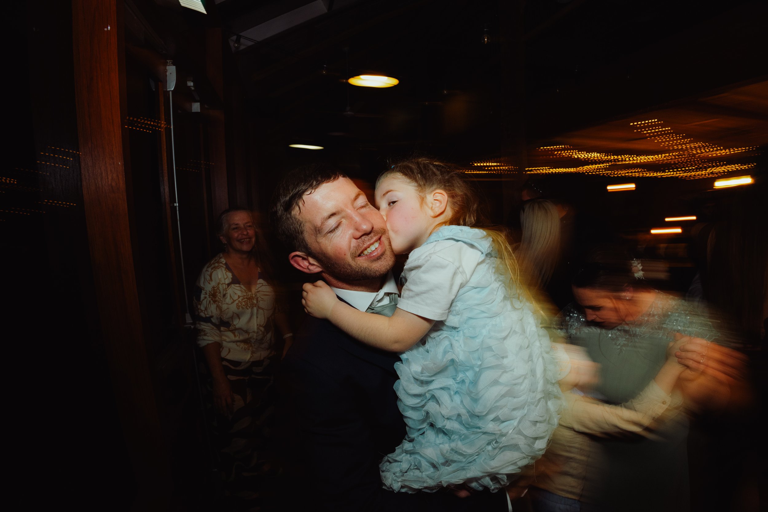 A man in a suit holding a young girl in a dress, kissing her cheek at a celebration or party with guests in the background.