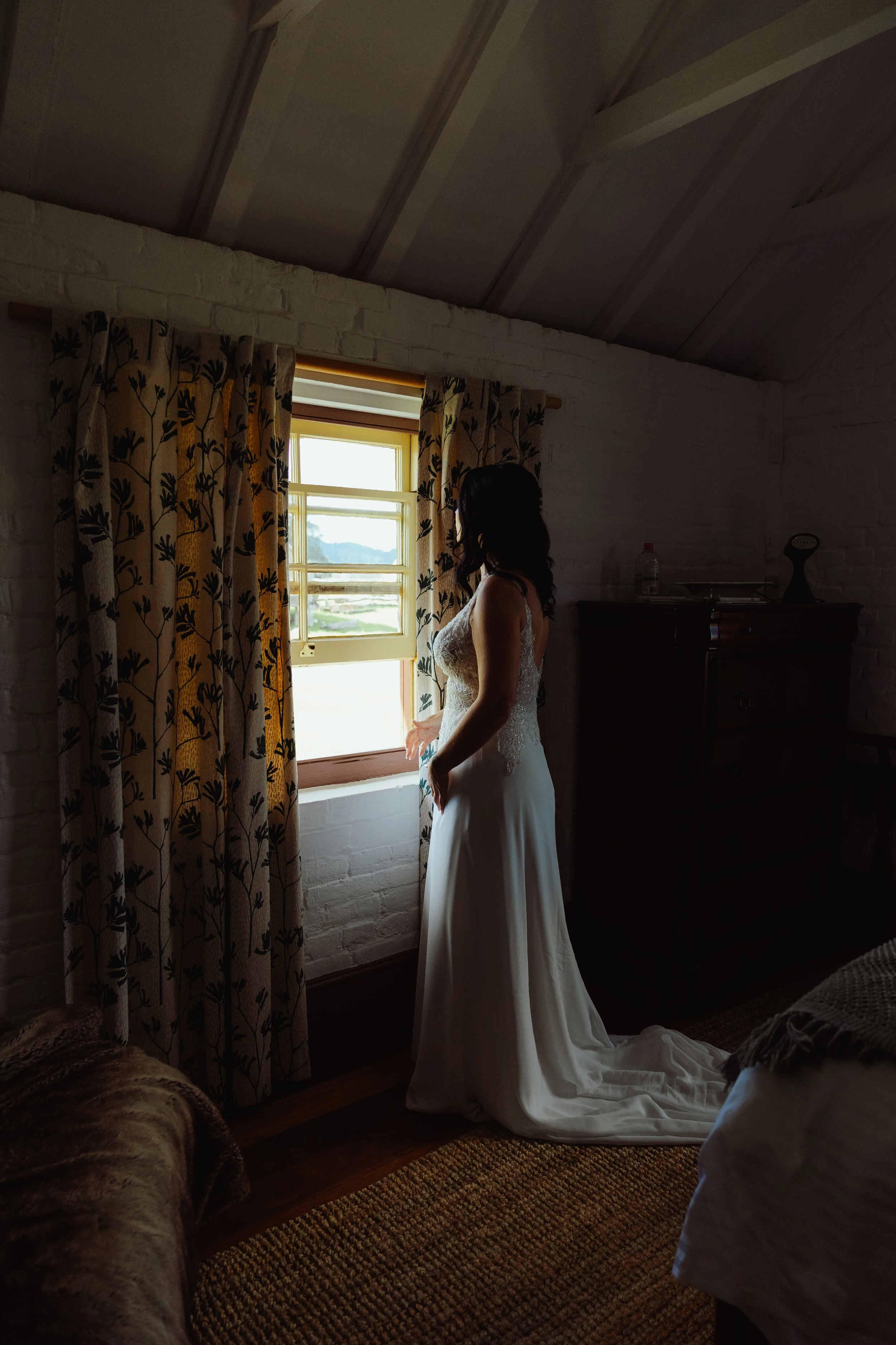 A woman in a wedding dress standing by a window in a cozy room with white brick walls. She is gazing outside, and sunlight is streaming through the window. The room has floral curtains and a dark cabinet with some objects on top.