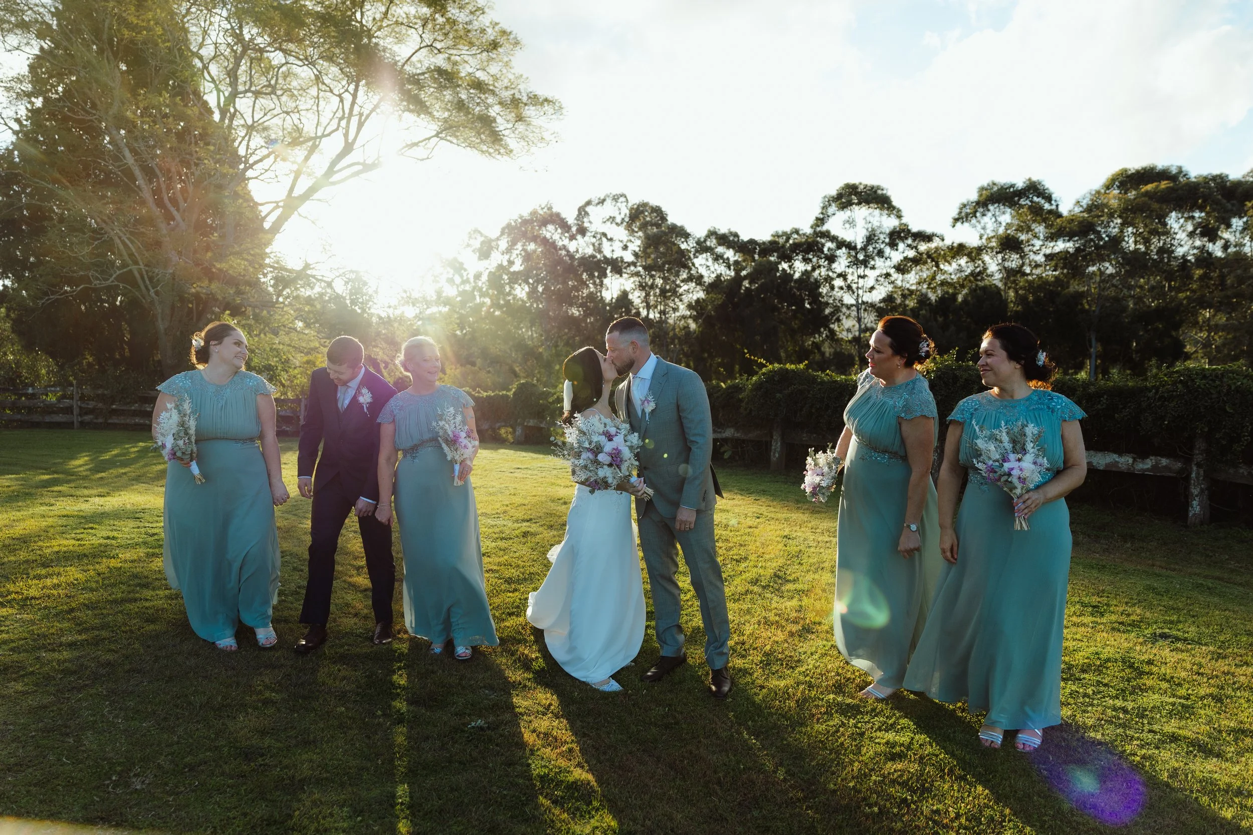 A wedding celebration outdoors with a bride and groom kissing among bridesmaids and groomsmen, under sunlight and trees.