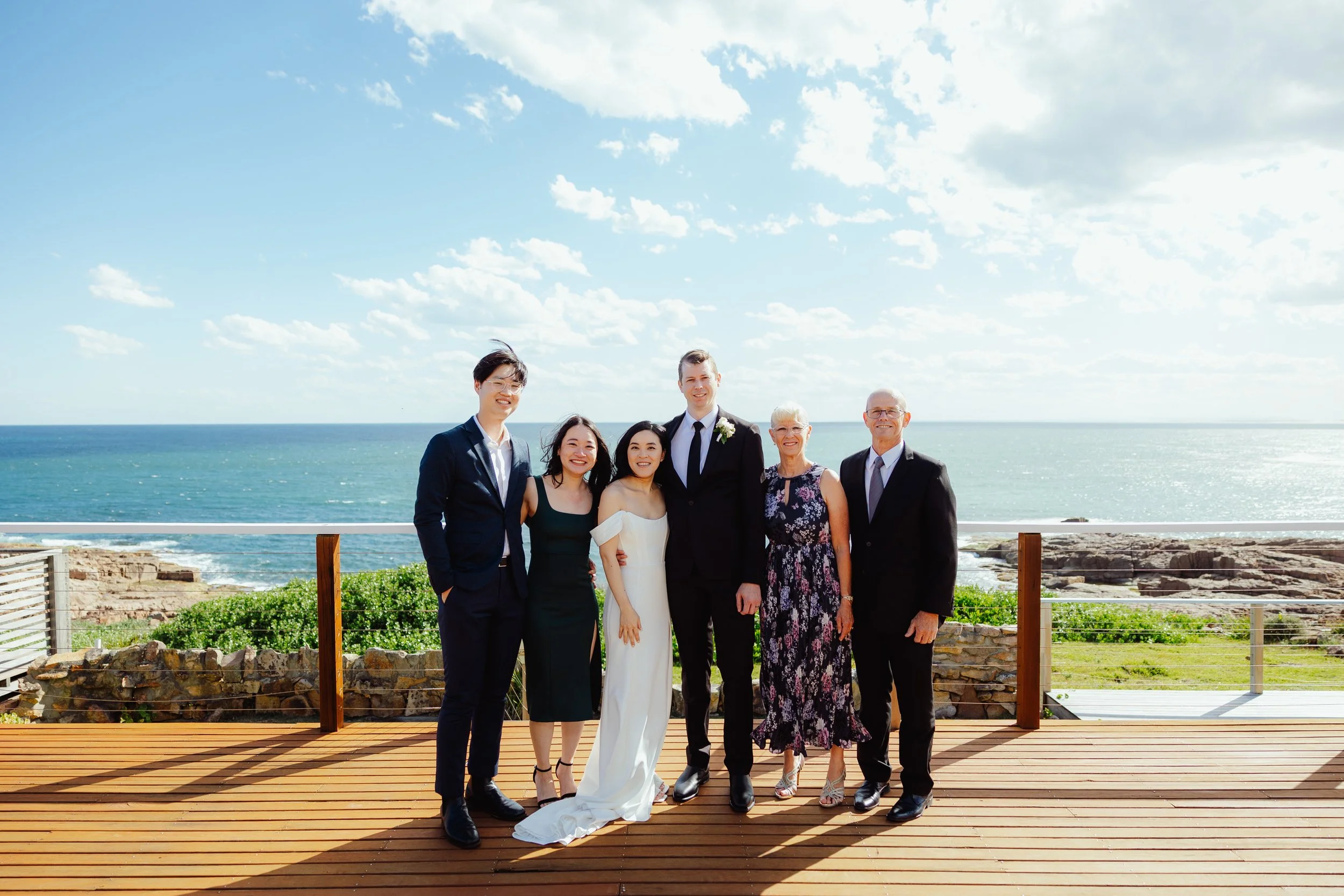 Group of seven people posing on a wooden deck near the ocean with a backdrop of blue sky and scattered clouds, with some rocky shoreline visible.