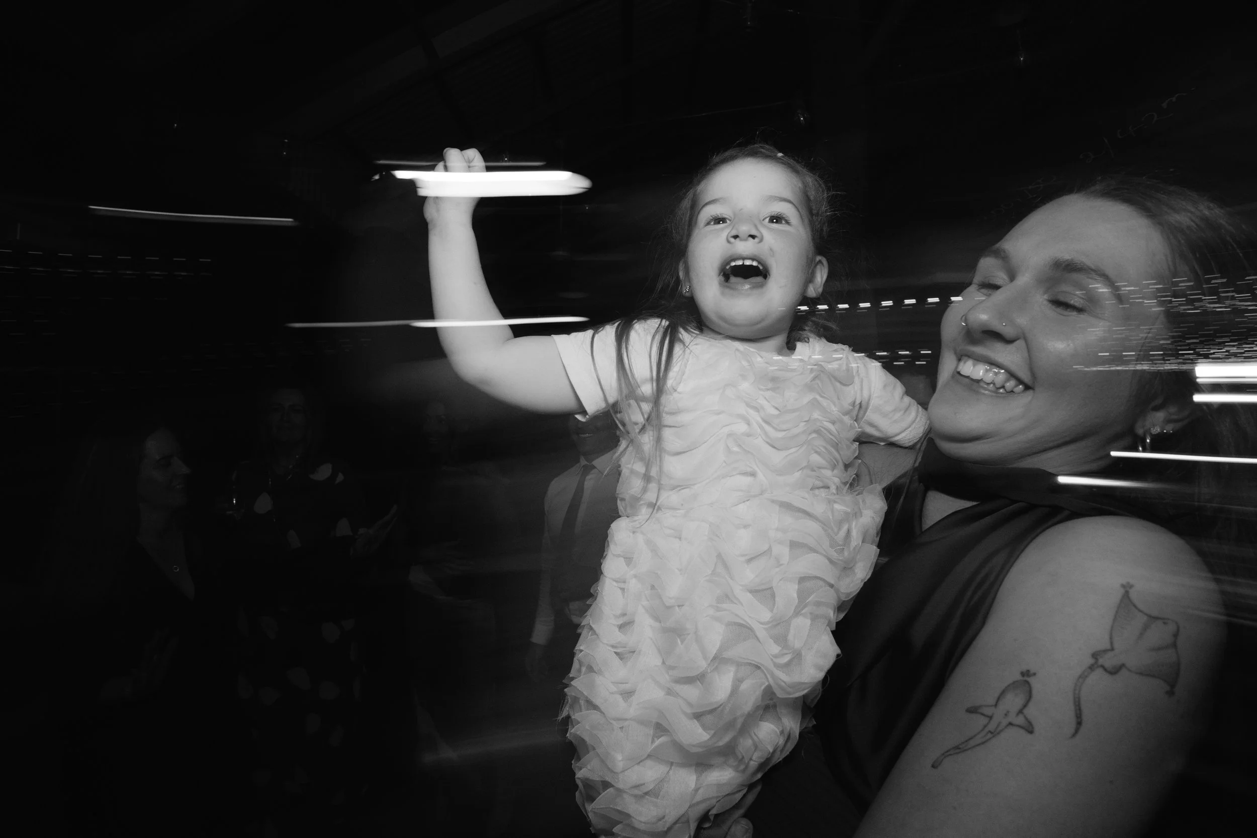 A woman lifts a young girl with her right hand. The girl has a joyful expression, her mouth open, eyes wide, and waves with her left hand. The woman has a big smile, eyes closed, and visible tattoos on her left arm. The image is in black and white.