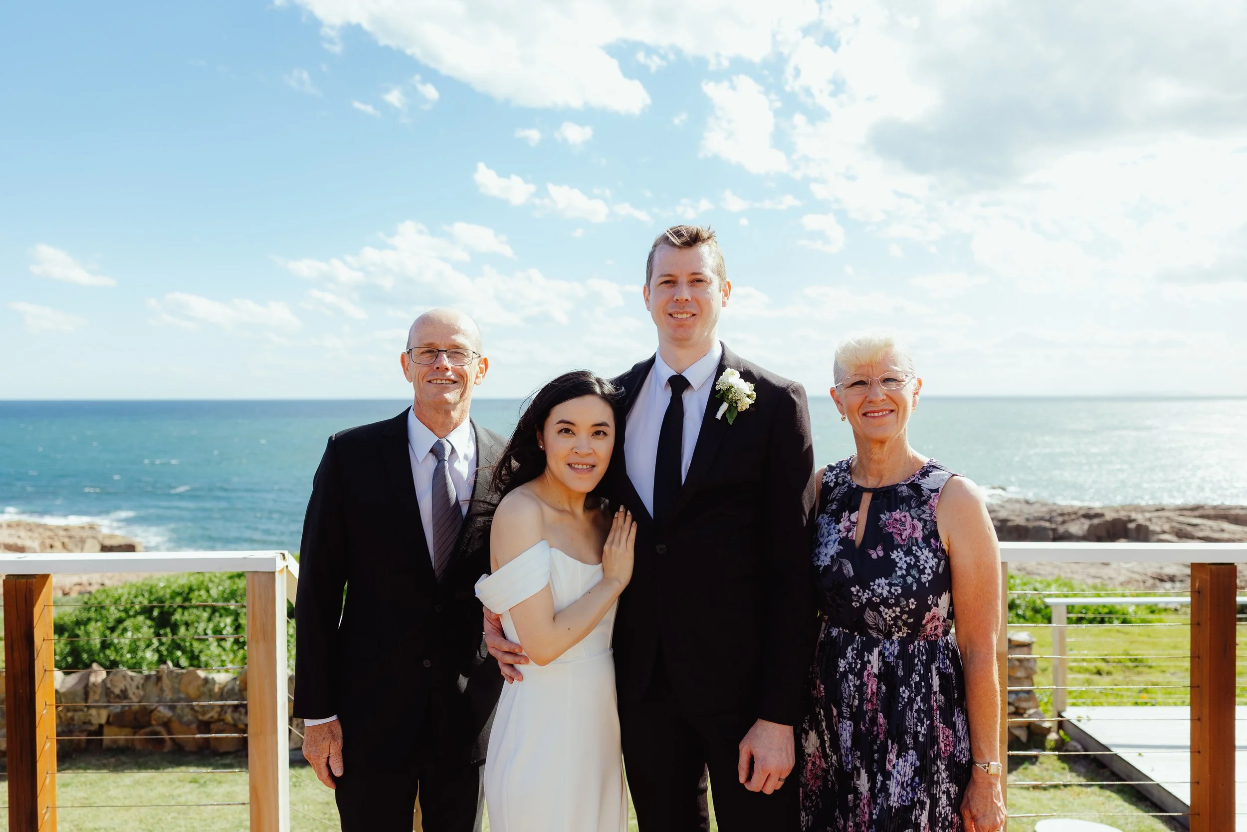 Group of five people, two men and three women, standing outdoors by the ocean on a sunny day, dressed in formal attire, at a wedding or special event.
