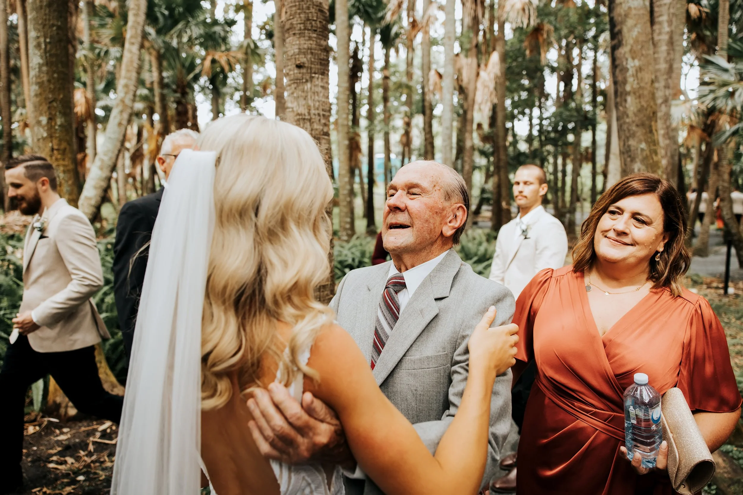 Bride and groom holding hands and looking at each other during a wedding ceremony outdoors, with wedding guests in the background among trees.