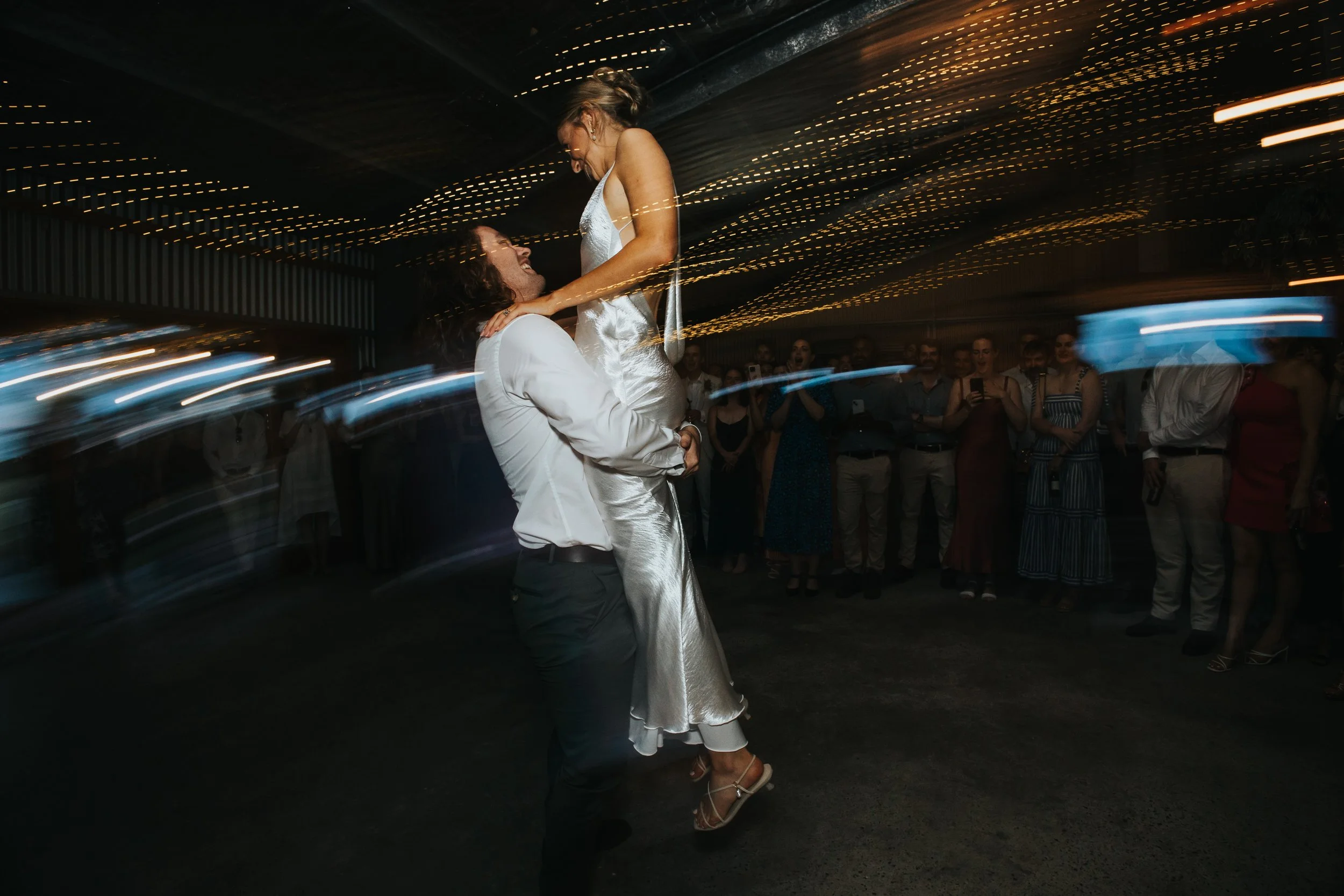 A bride and groom are dancing closely at their wedding reception, surrounded by guests, under a ceiling decorated with string lights. The bride is wearing a satin wedding gown and heels, and the groom is dressed in a white suit jacket. The couple is 