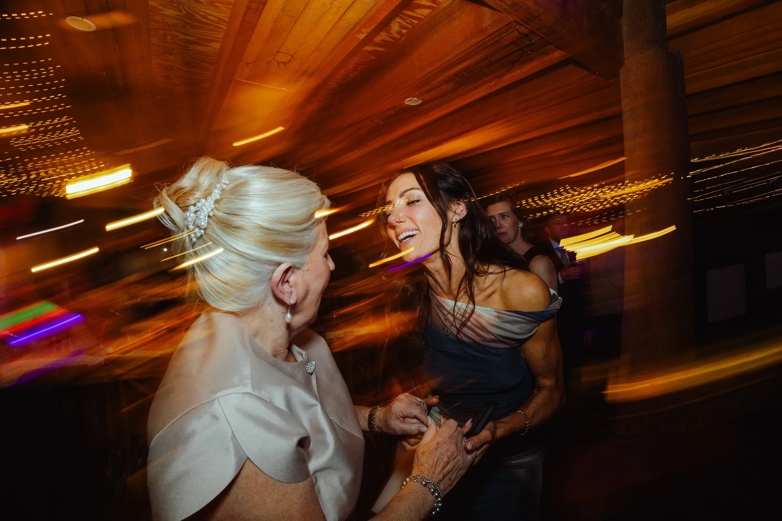 Two women dancing and smiling at a party, with colorful lights and a blurred background.