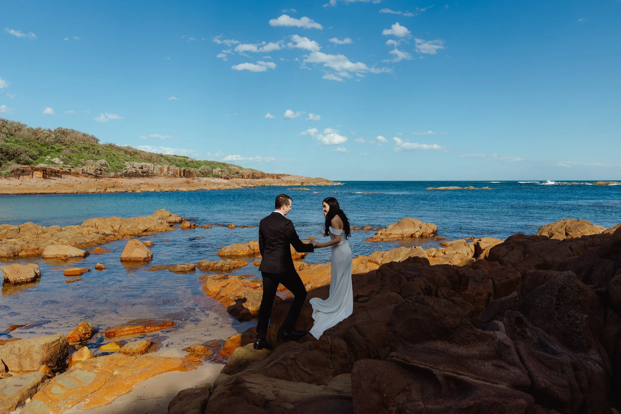 A man and a woman dressed in formal wedding attire holding hands on a rocky beach with the ocean behind them and a clear blue sky with scattered clouds.