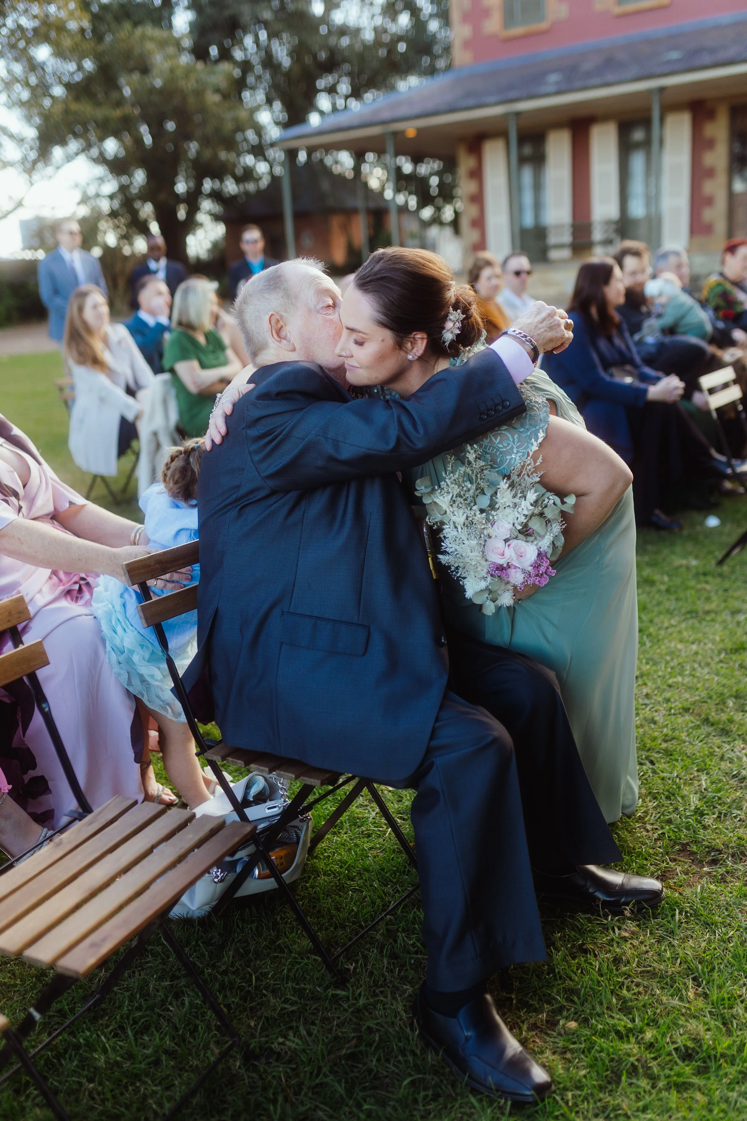 An elderly man in a suit embraces a woman in a light green dress at an outdoor wedding ceremony. Guests seated on wooden chairs are visible in the background near a house with a porch, and some guests are wearing sunglasses.
