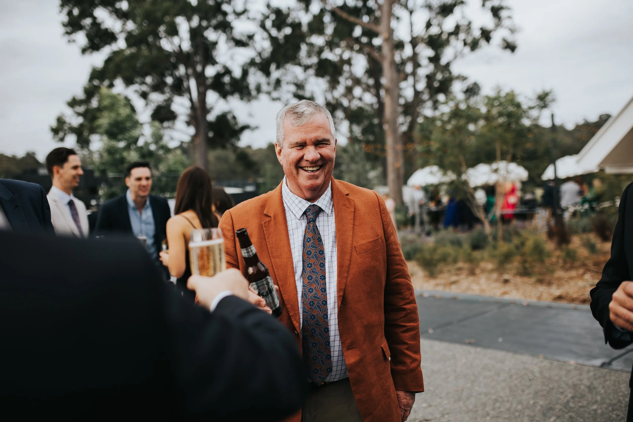 A group of people at an outdoor gathering, with a man in an orange jacket smiling and holding a bottle of beer, while others in the background are engaged in conversation.