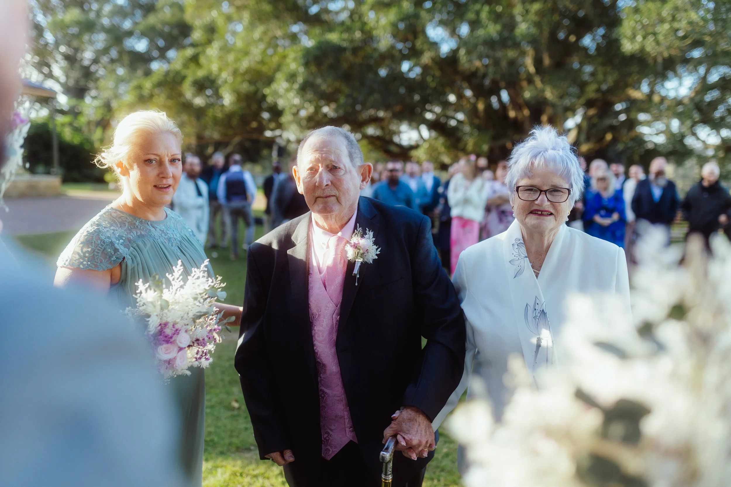 Three elderly people, two women and one man, standing outdoors during a social gathering or wedding, with a background of trees and other guests, the man holding a cane and wearing a dark suit with a pink vest and boutonniere.