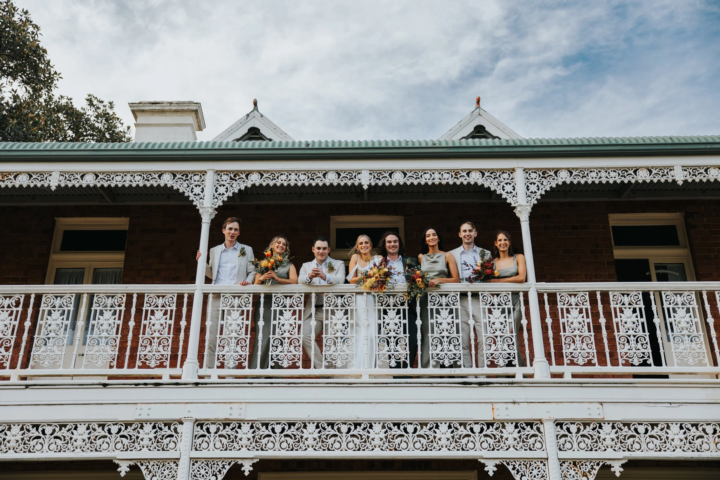 A group of people dressed in formal attire standing on a balcony with intricate white wrought iron railings, with a brick building in the background.