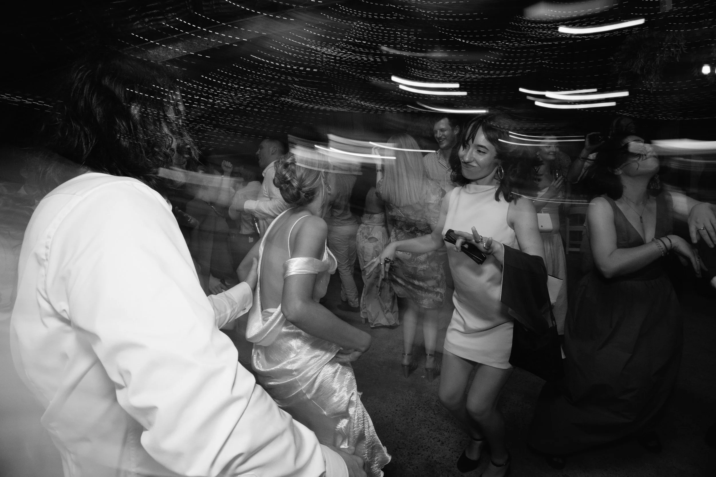 Black and white photo of people dancing and socializing in a lively indoor setting with string lights.