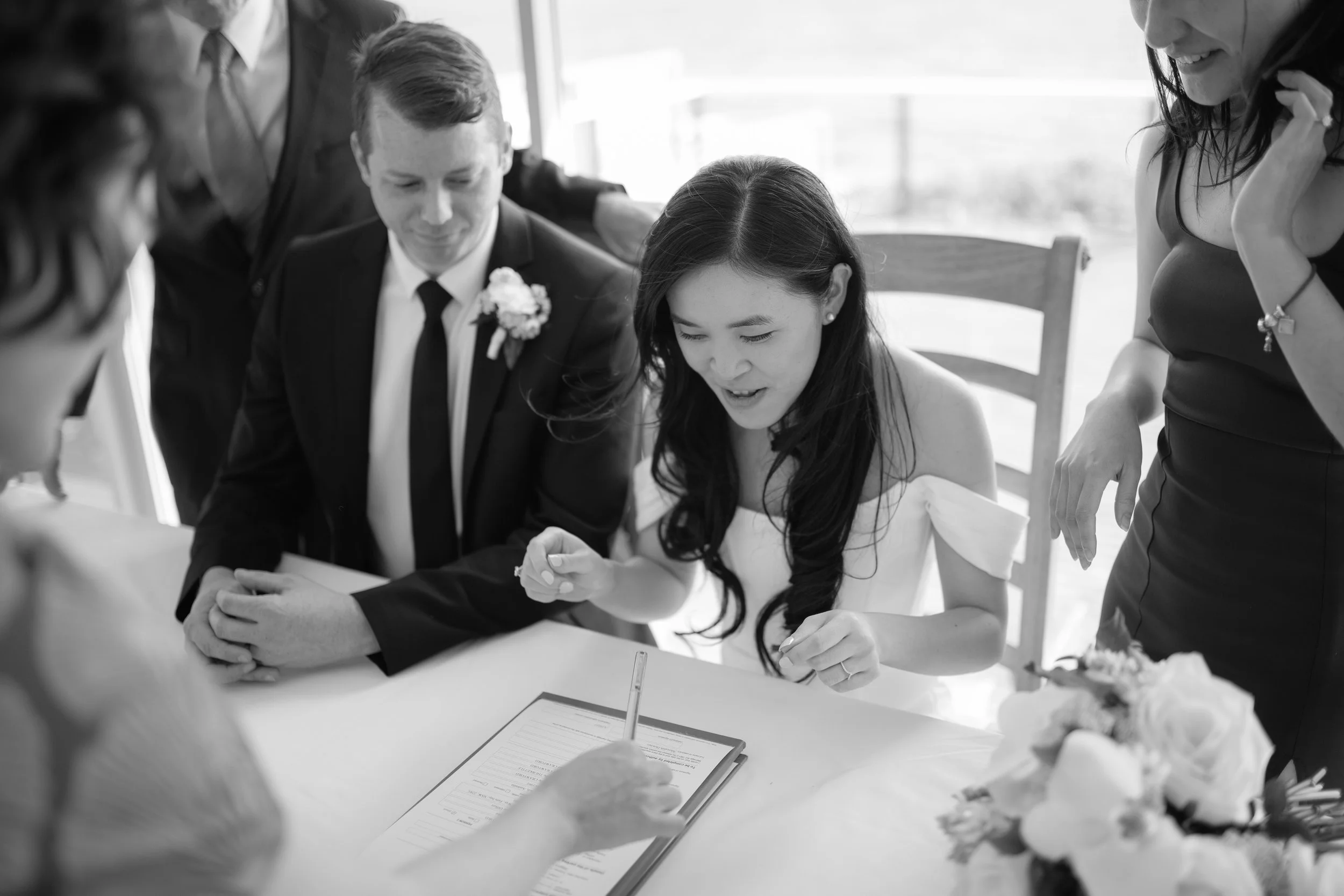 A wedding ceremony where a bride signs a document on a table while surrounded by people. The groom is sitting next to her, dressed in a suit with a boutonniere. The bride wears a white dress and has long dark hair. Several women are standing nearby, 