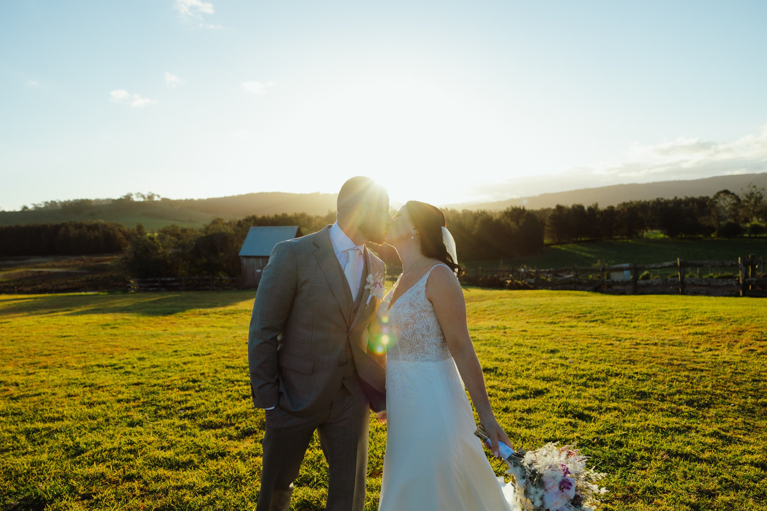 A bride and groom kissing outdoors at sunset on grass, with a barn and hills in the background.