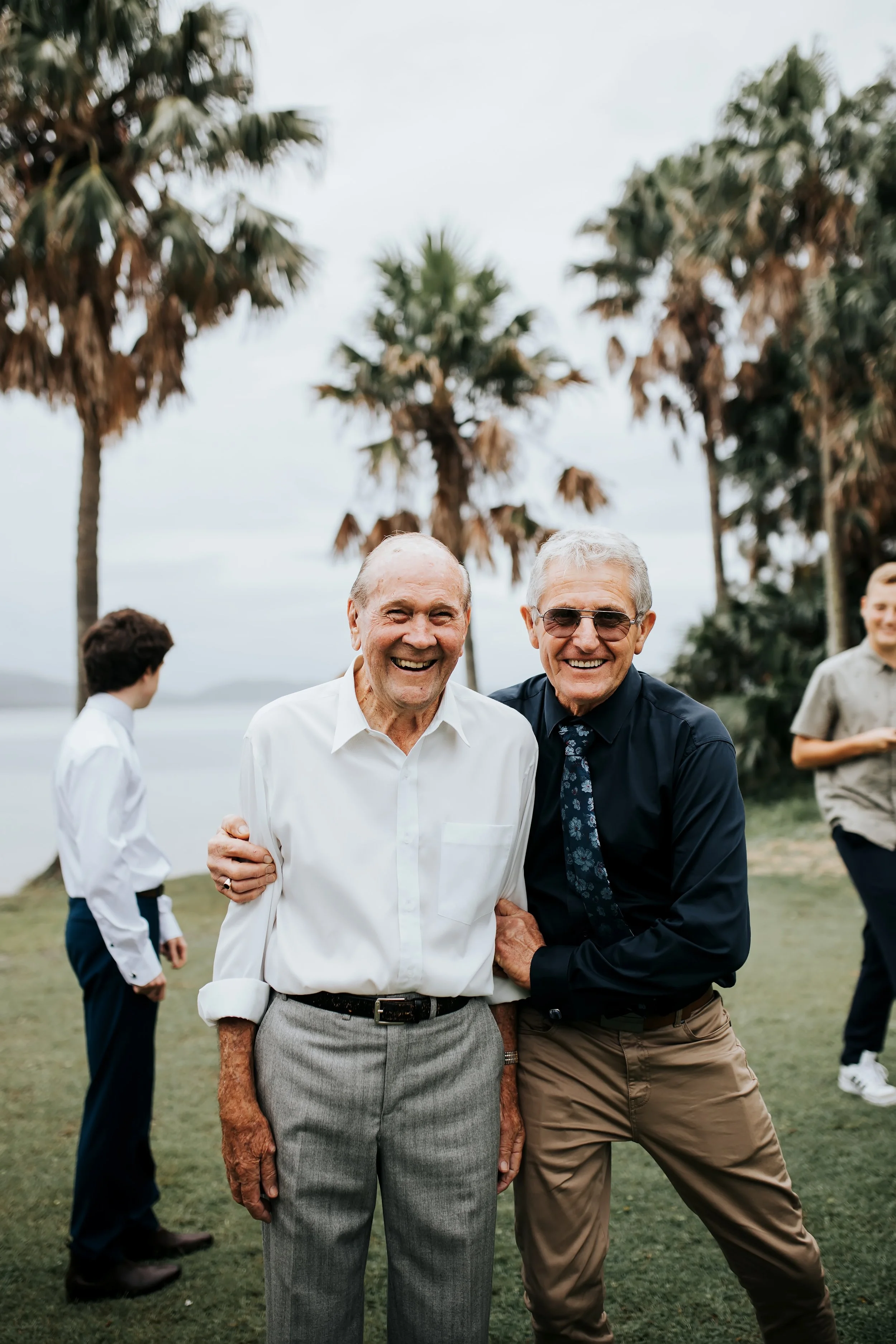 Two elderly men smiling and holding hands outdoors with palm trees in the background, celebrating together.