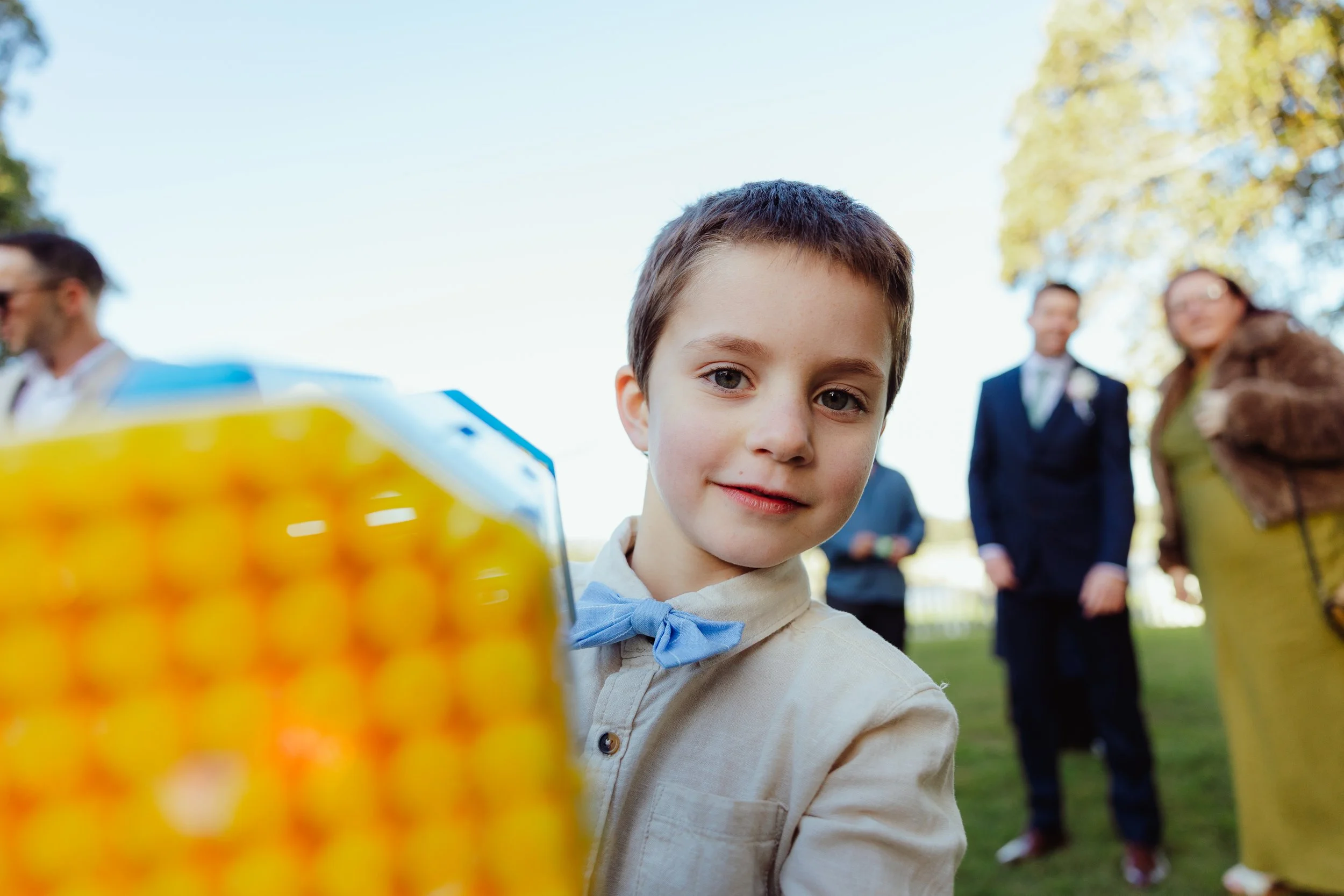 Close-up of a young boy with short brown hair and blue eyes wearing a white shirt with a blue bow tie, outdoors on a sunny day with trees in the background, surrounded by adults who are blurred in the background.
