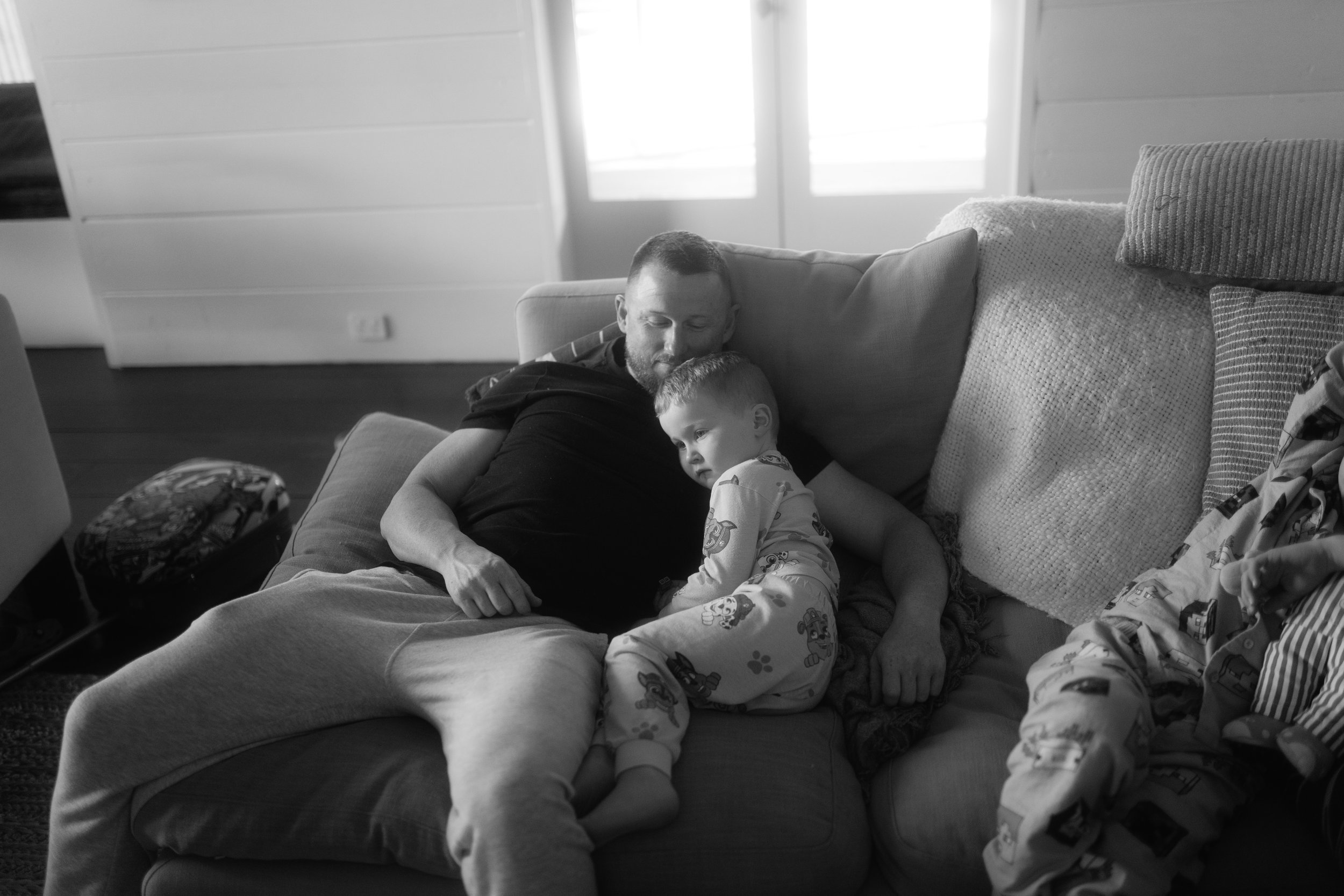A man and a young child sitting on a couch together, appearing to be resting or napping. The man has a beard and is dressed casually, while the child is wearing pajamas with cartoon characters. The setting is a cozy living room with pillows and a win