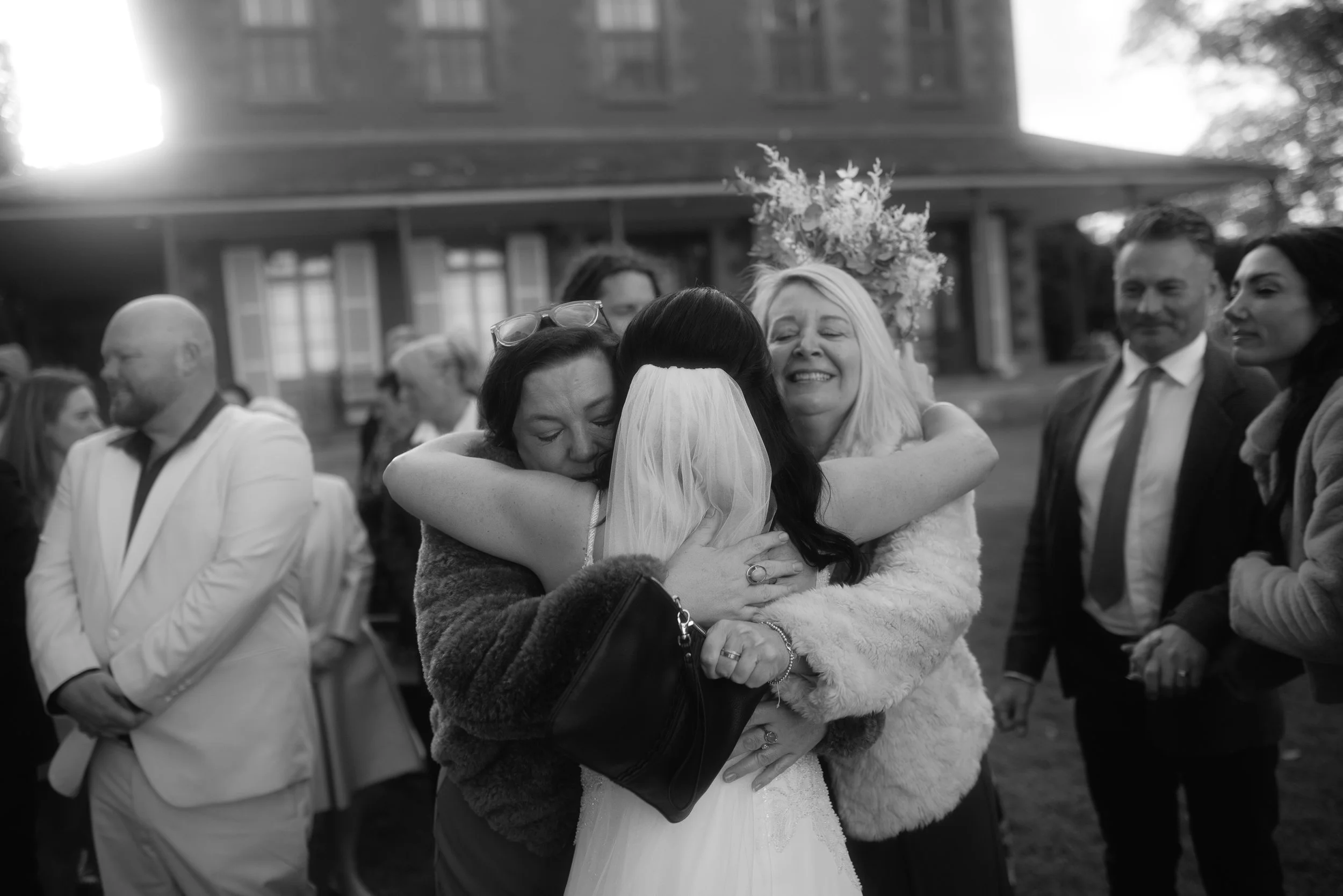 Black and white photo of people at a wedding, with two women hugging in the center, a woman with a floral crown, and several guests standing nearby.