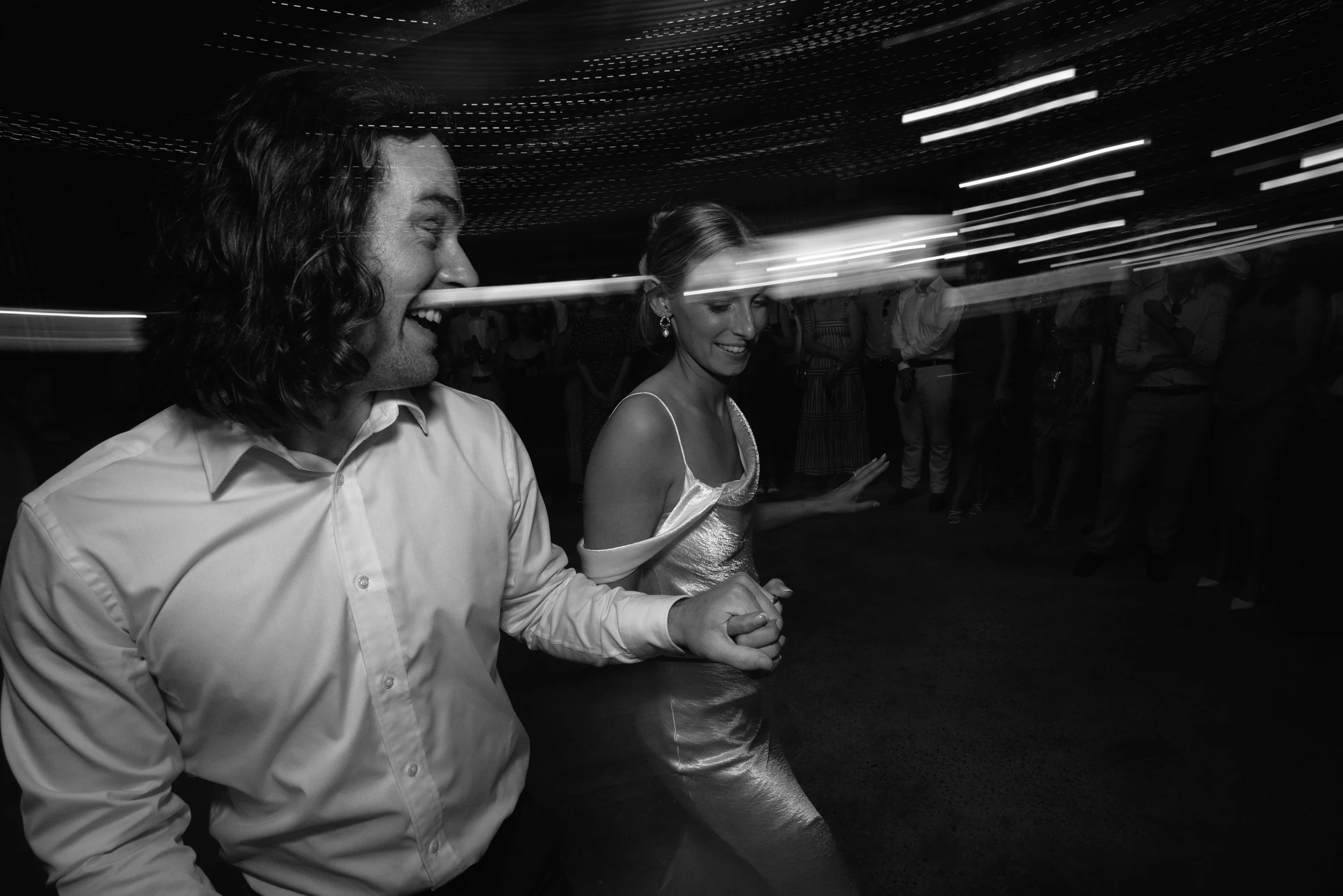 A black and white photo of a man and woman dancing together at a party or celebration, surrounded by other guests, with light streaks indicating motion or dancing.