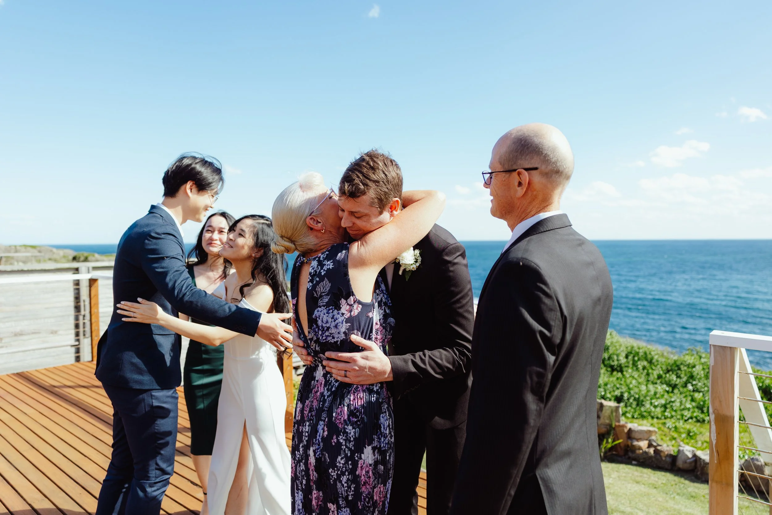 A group of six people, including two men and four women, dressed in formal attire, are embracing and smiling during a wedding ceremony outdoors near the ocean on a wooden deck, with a blue sky and few clouds in the background.